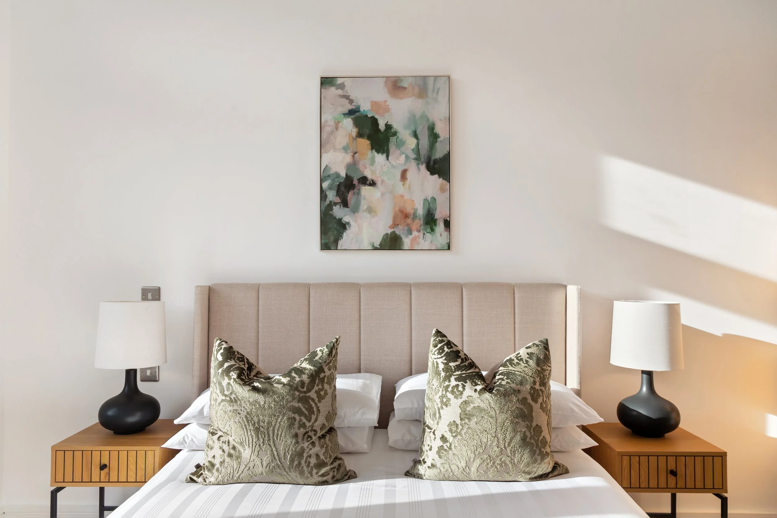 A bedroom with a beige upholstered headboard, two matching black and white lamps on wooden side tables, two decorative pillows with a gold and cream damask pattern, white bedding, and an abstract painting on the white wall above the headboard.