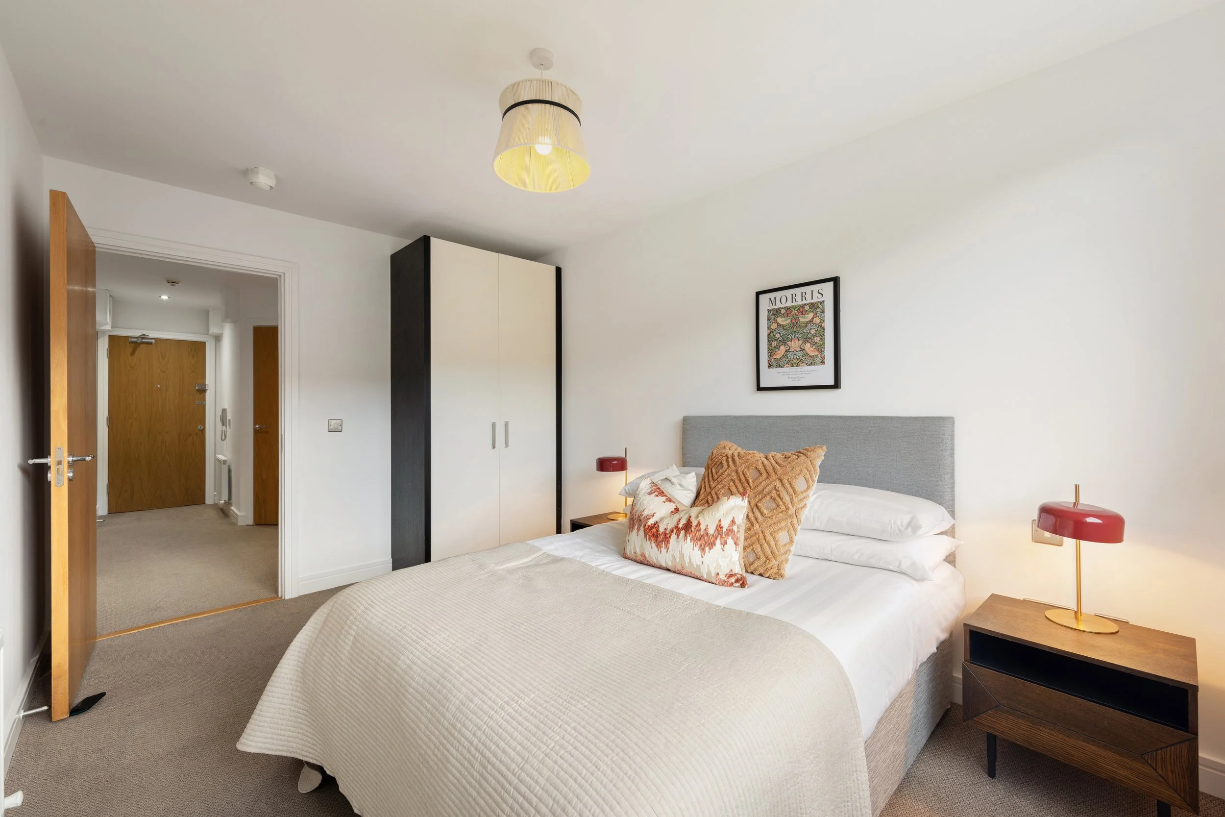 A cozy bedroom featuring a gray upholstered bed with decorative pillows, wooden nightstand with a red lamp, a black and white wardrobe, and a framed artwork on the wall.
