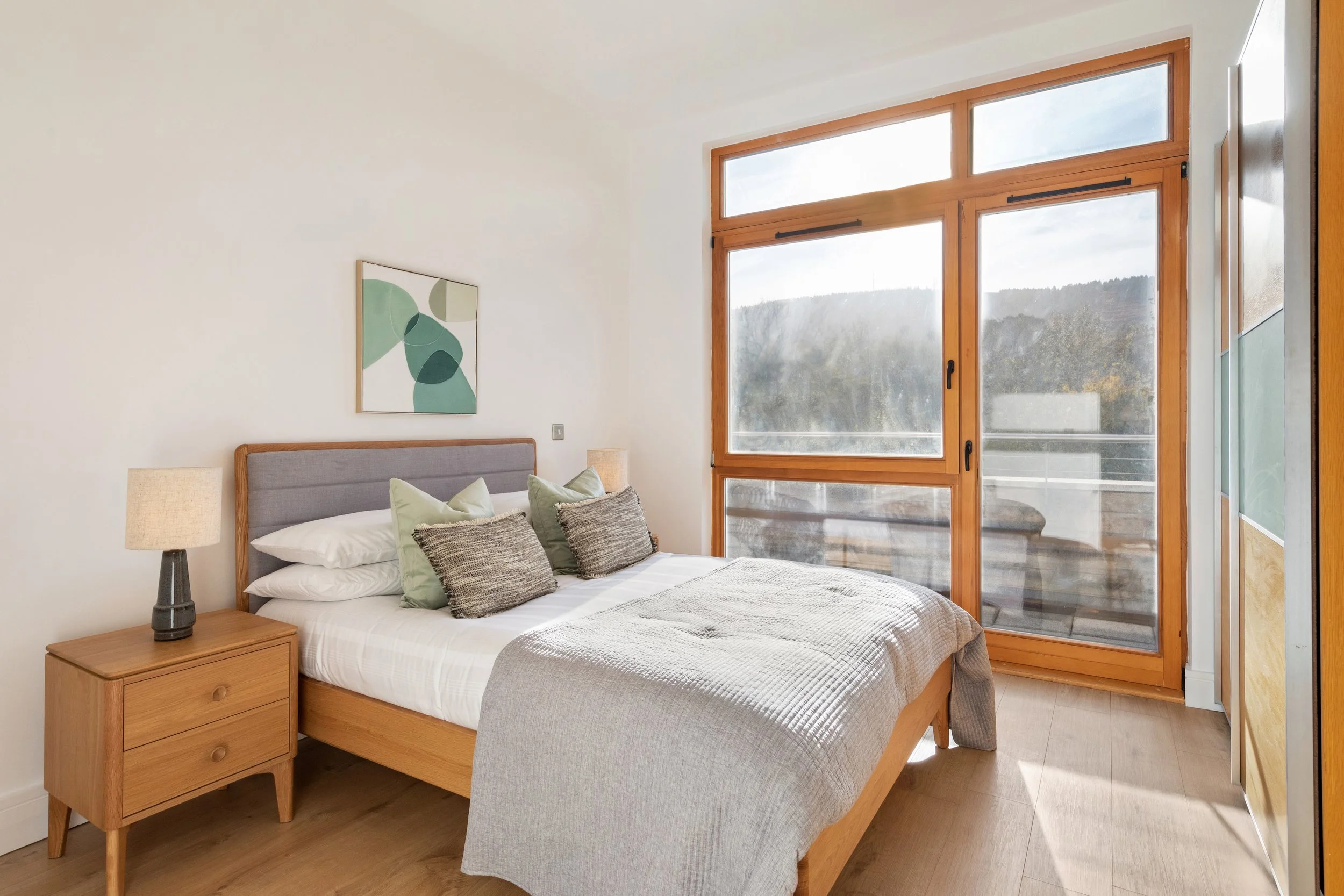 A bedroom with a bed, nightstand, lamp, and large window showing an outdoor view with trees and mountains.