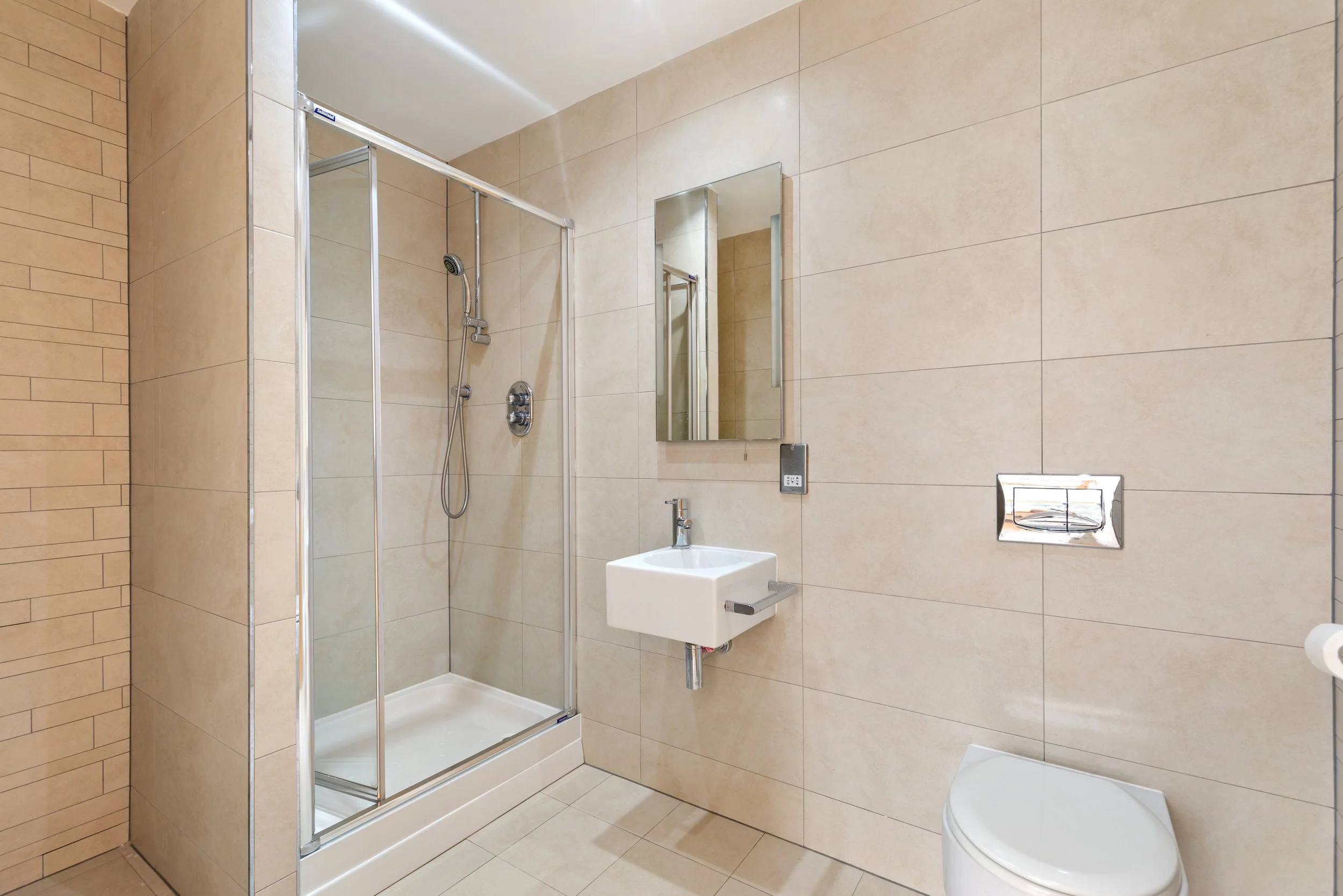 Modern bathroom with beige tiled walls and floor, clear glass shower enclosure, wall-mounted sink, mirror, toilet, and electronic control on wall