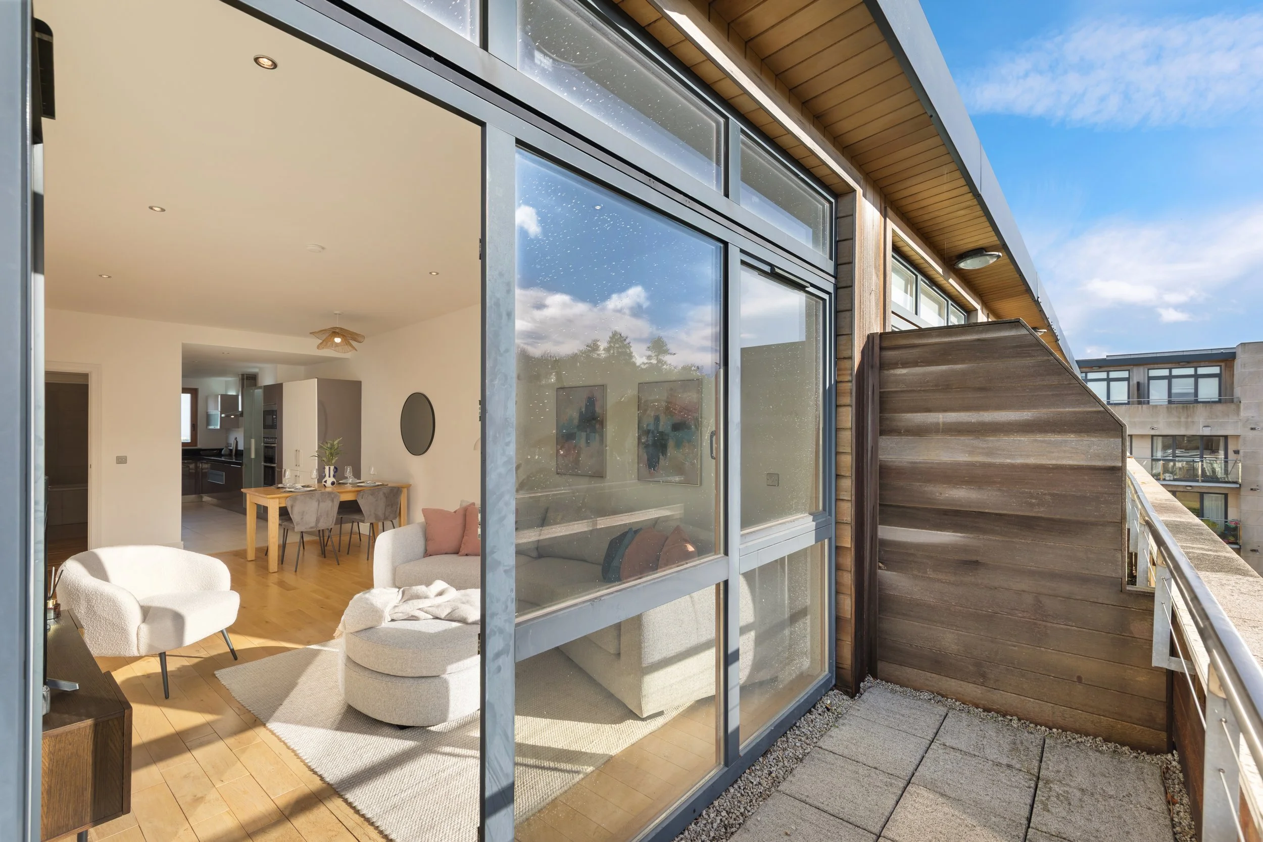 View of an apartment balcony with glass sliding doors, showing a modern living room with white walls, wooden flooring, a beige sofa with pink pillows, a white armchair, and a dining area with a wooden table and gray chairs. The balcony has a wooden p
