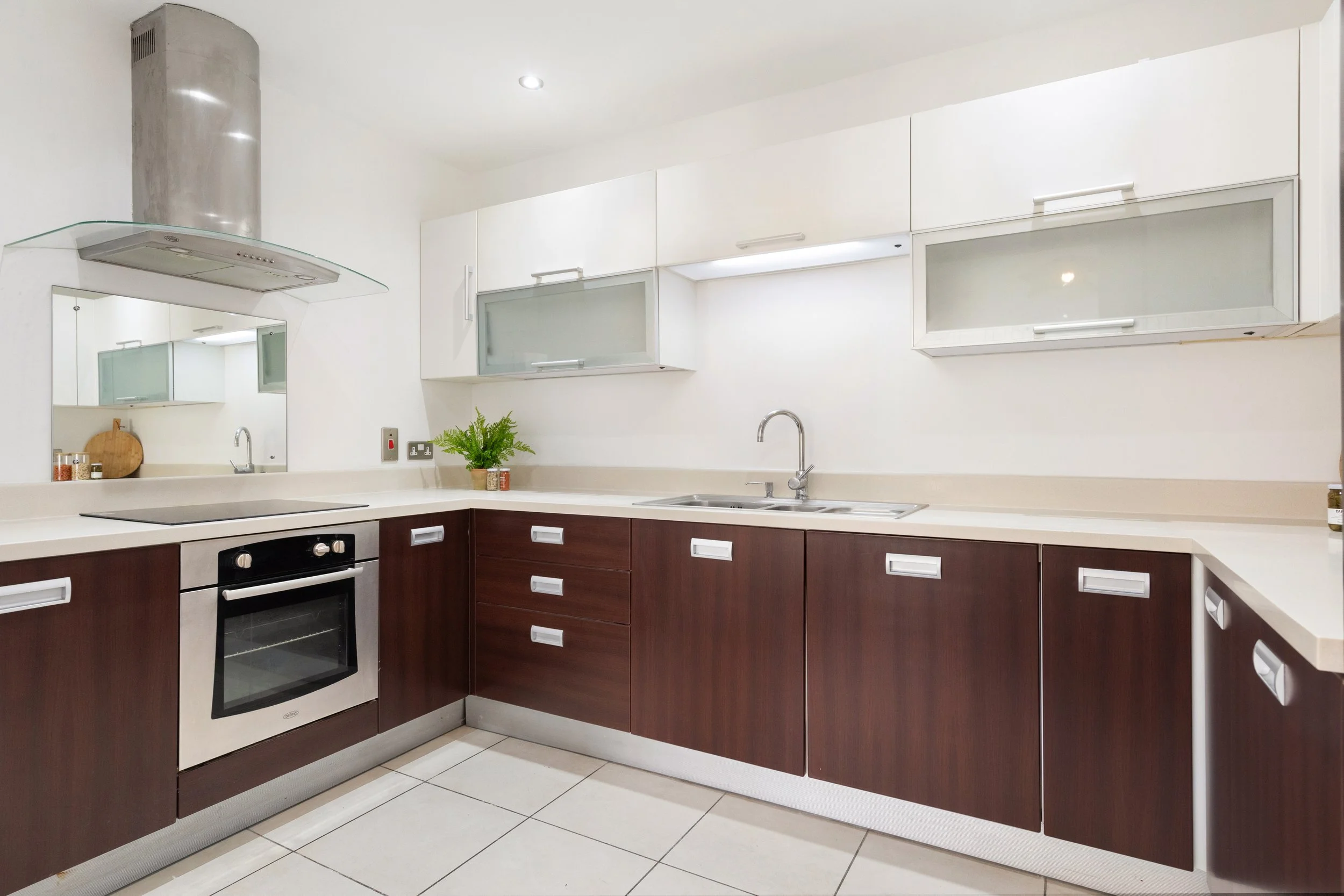 Modern kitchen with white upper cabinets, dark wood lower cabinets, stainless steel oven, and sink with faucet. Light-colored countertops, a small green plant, and a mirror on the wall.