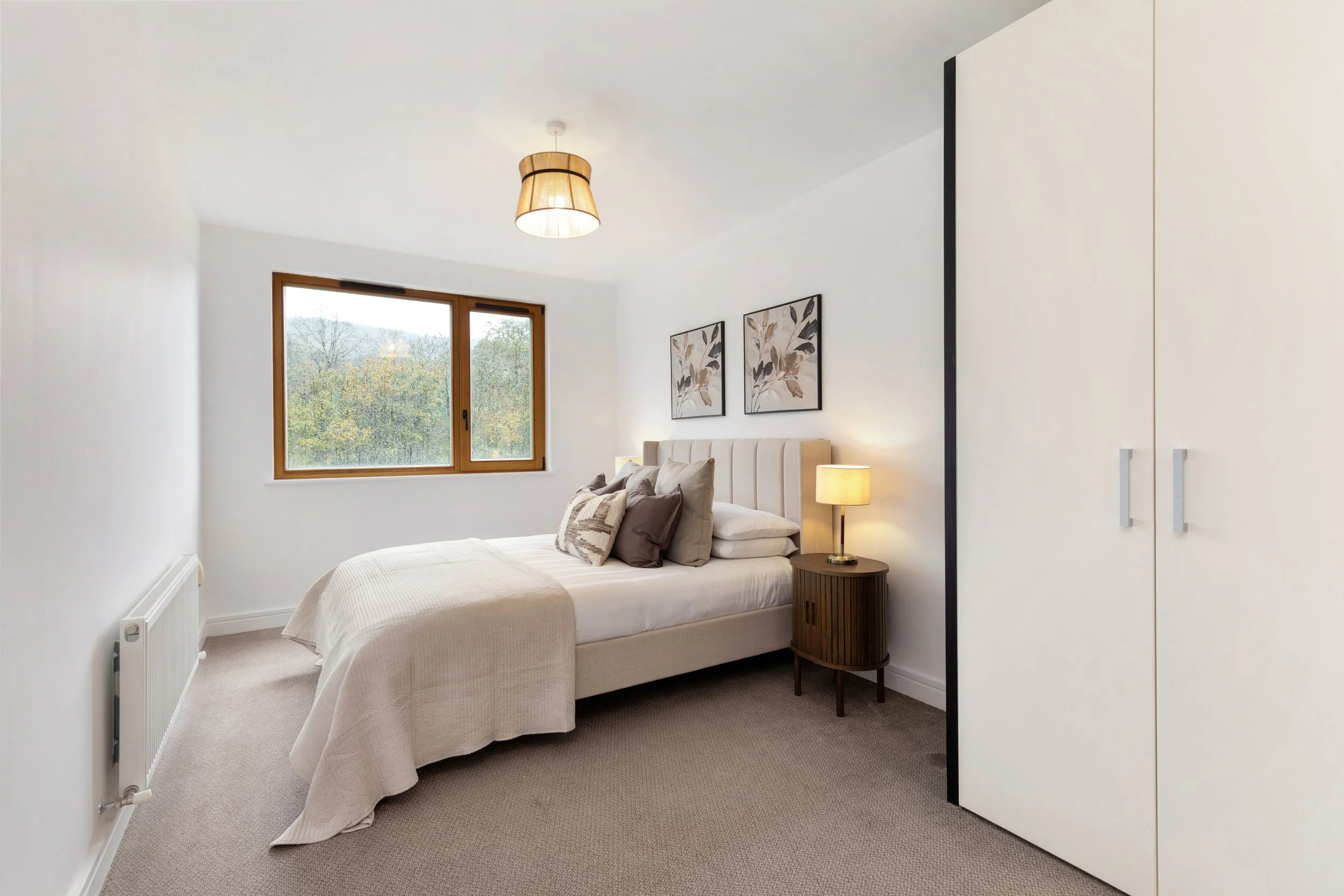 A bedroom with a bed, nightstand, lamp, framed artwork, window, and closet in neutral tones.