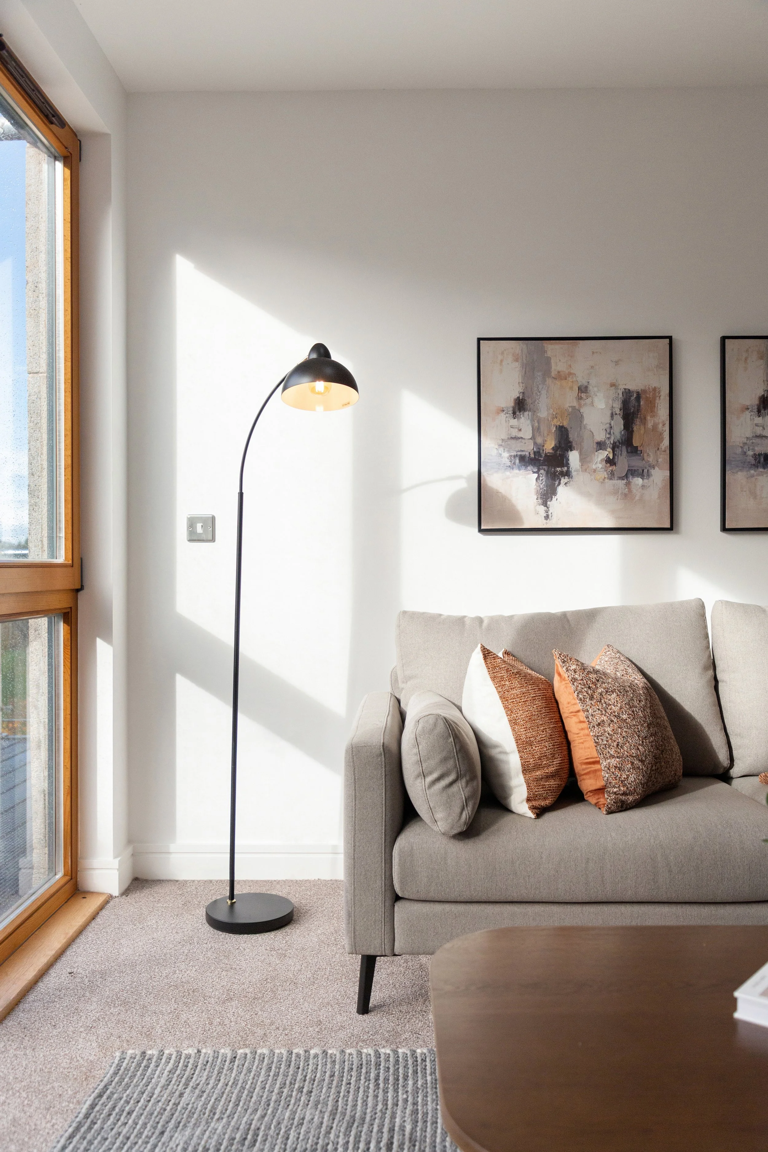 A cozy living room corner with a beige sofa decorated with various brown and orange pillows, a black floor lamp, a wooden window with natural light streaming in, abstract wall art, a textured area rug, and part of a wooden coffee table.
