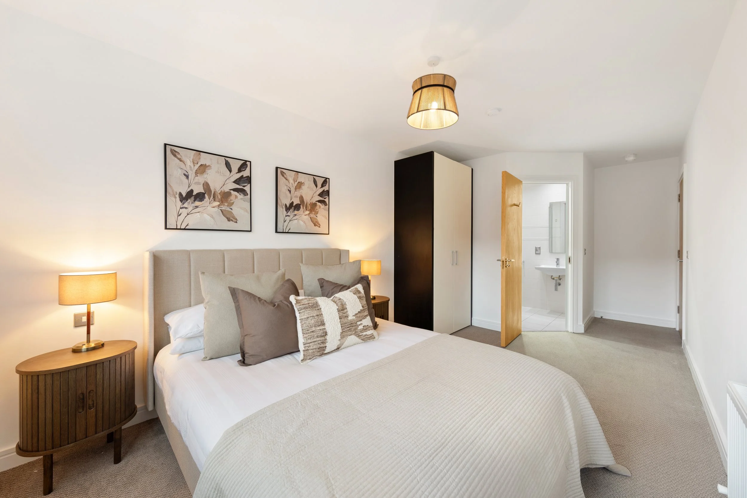 Bedroom with a bed, nightstand, lamps, wall art, wardrobe, and bathroom visible through open door.