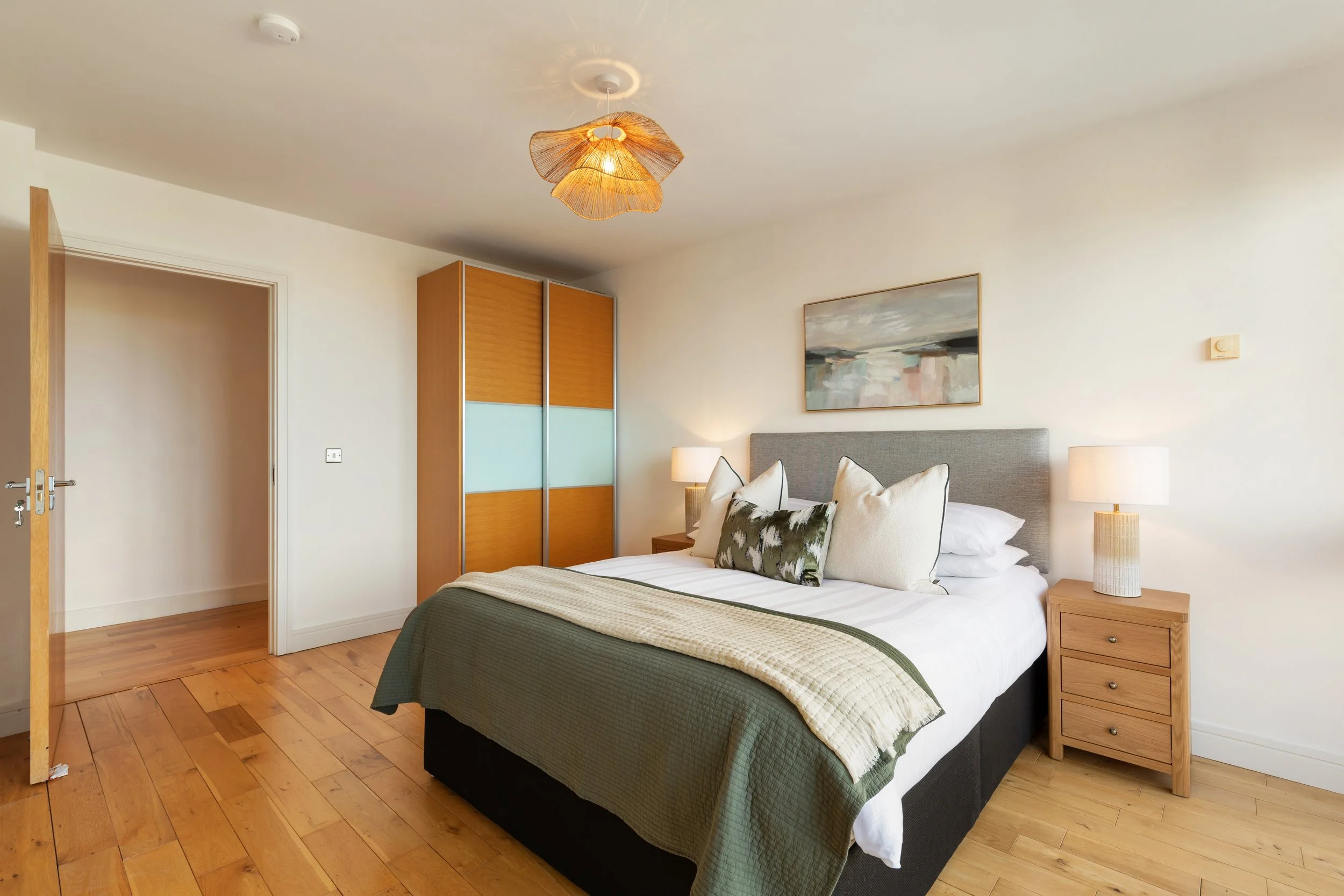 A neatly made bedroom with a bed, side tables, lamps, a painting, and a wardrobe with sliding doors.