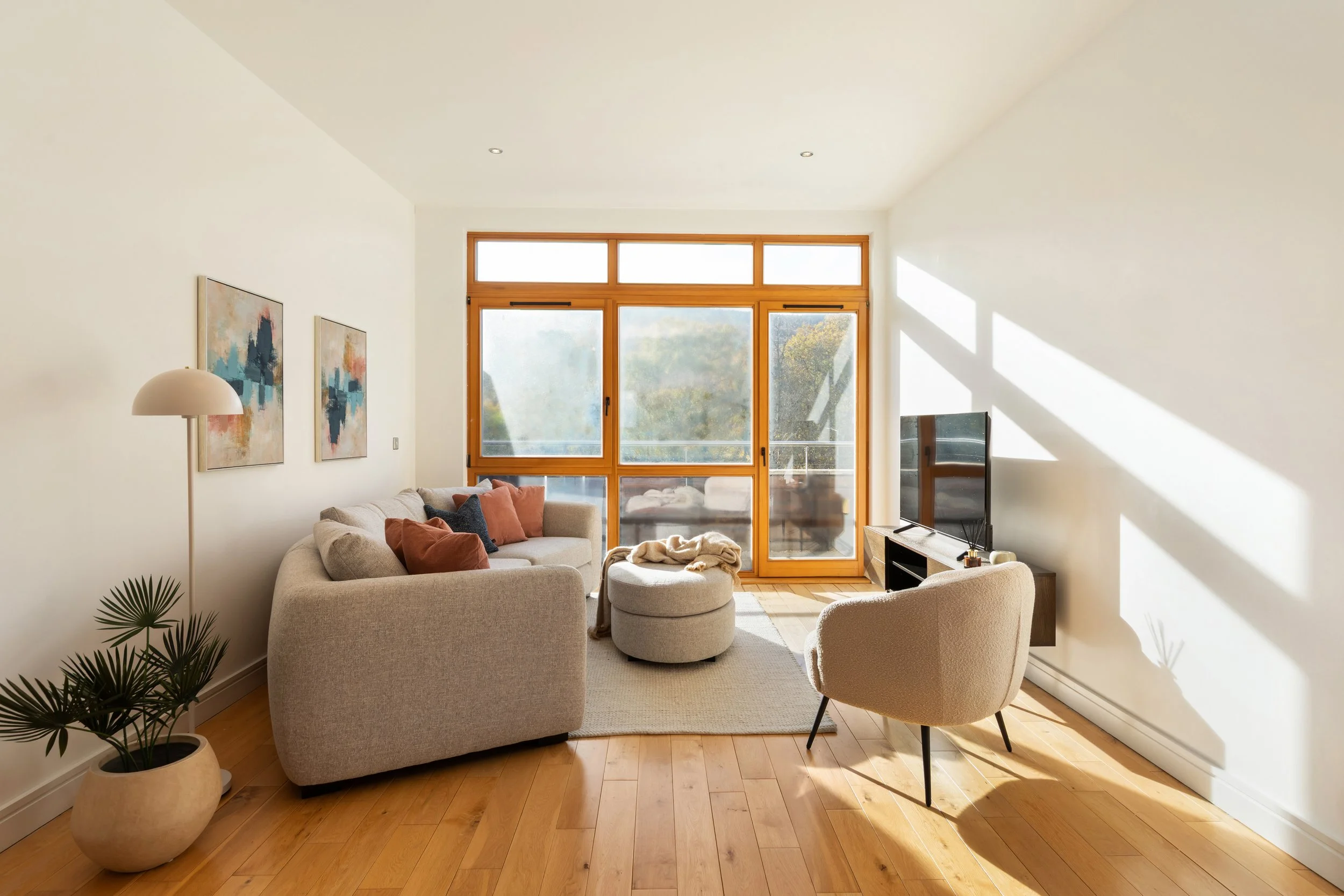 Bright living room with a beige sofa with throw pillows, a white armchair, a small ottoman with a blanket, a wooden TV stand with a flat-screen TV, and large windows letting in sunlight, with hardwood flooring and minimal decor.