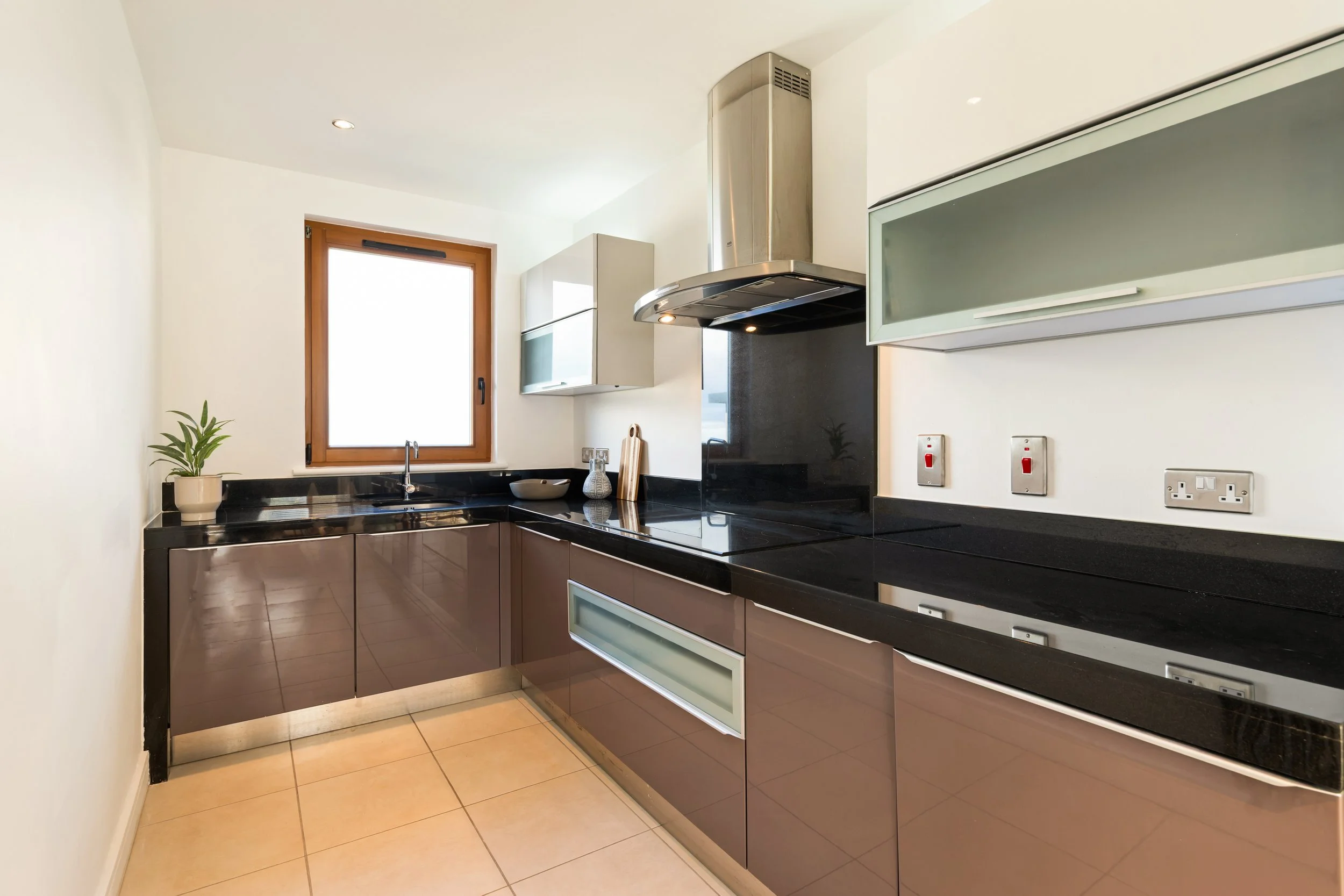 Modern kitchen with black countertops, brown cabinetry, a stainless steel range hood, and a window above the sink, decorated with a potted plant and small decorative items.