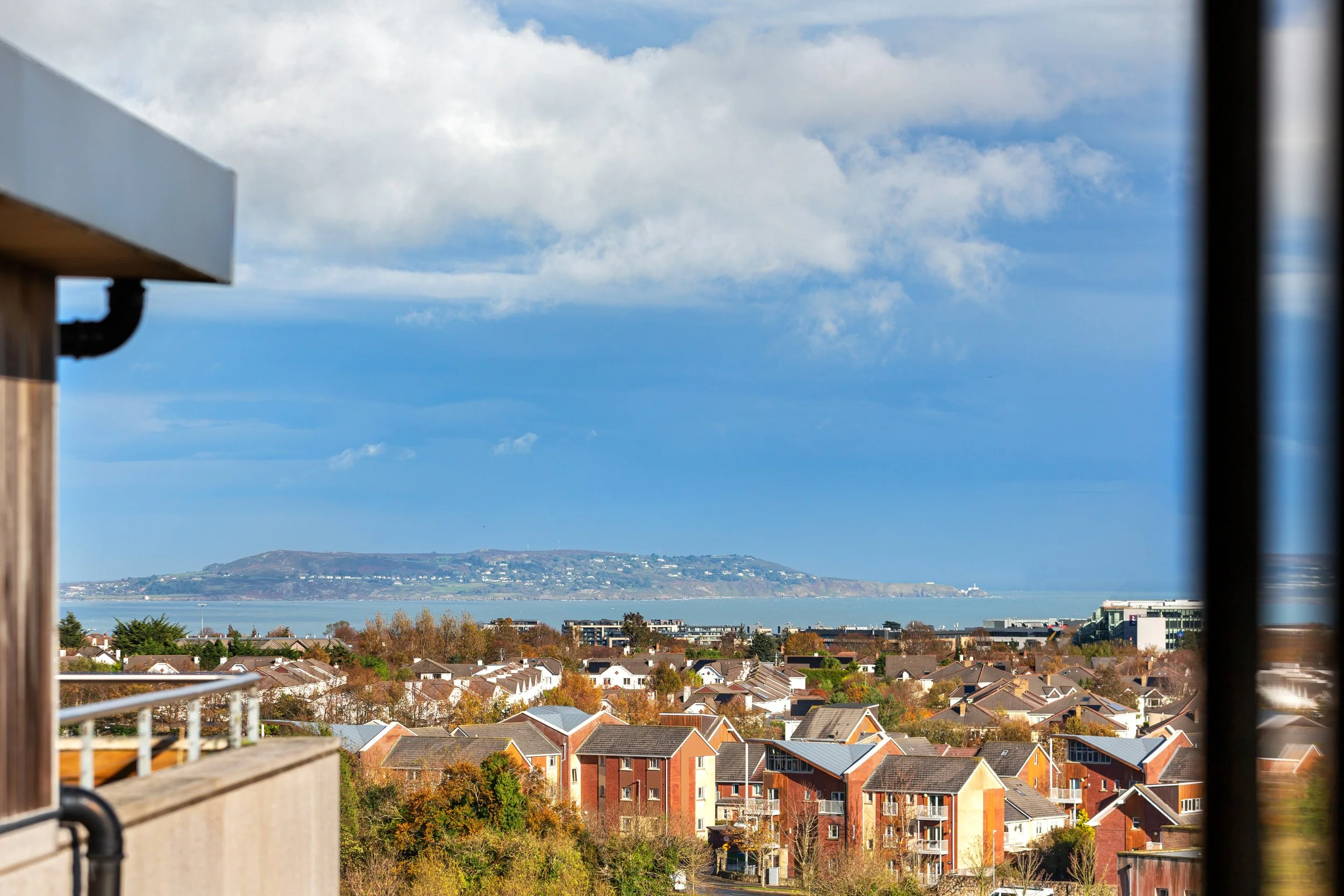 Scenic view of a residential area with houses, trees, and a body of water in the distance, with a hill or island on the horizon under a partly cloudy sky.