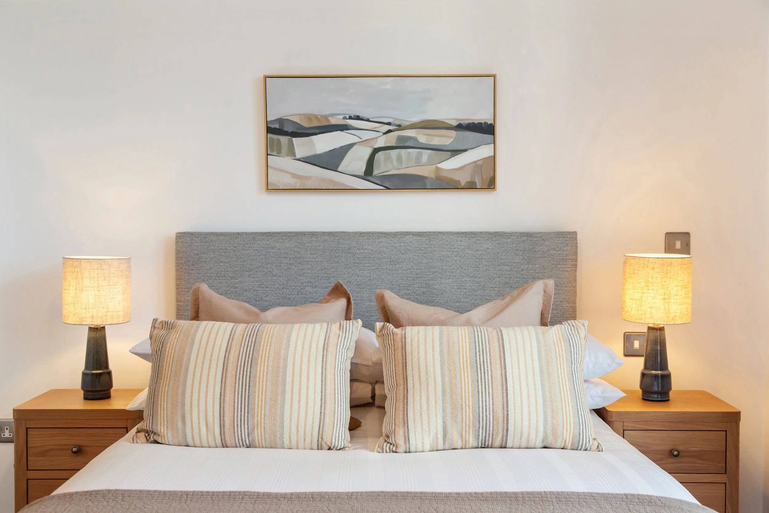 A nicely made bed with a gray upholstered headboard, four pillows with beige pillowcases, and two striped pillows in front. On each side of the bed are wooden nightstands with matching lamps with beige lampshades. A framed landscape painting hangs ab