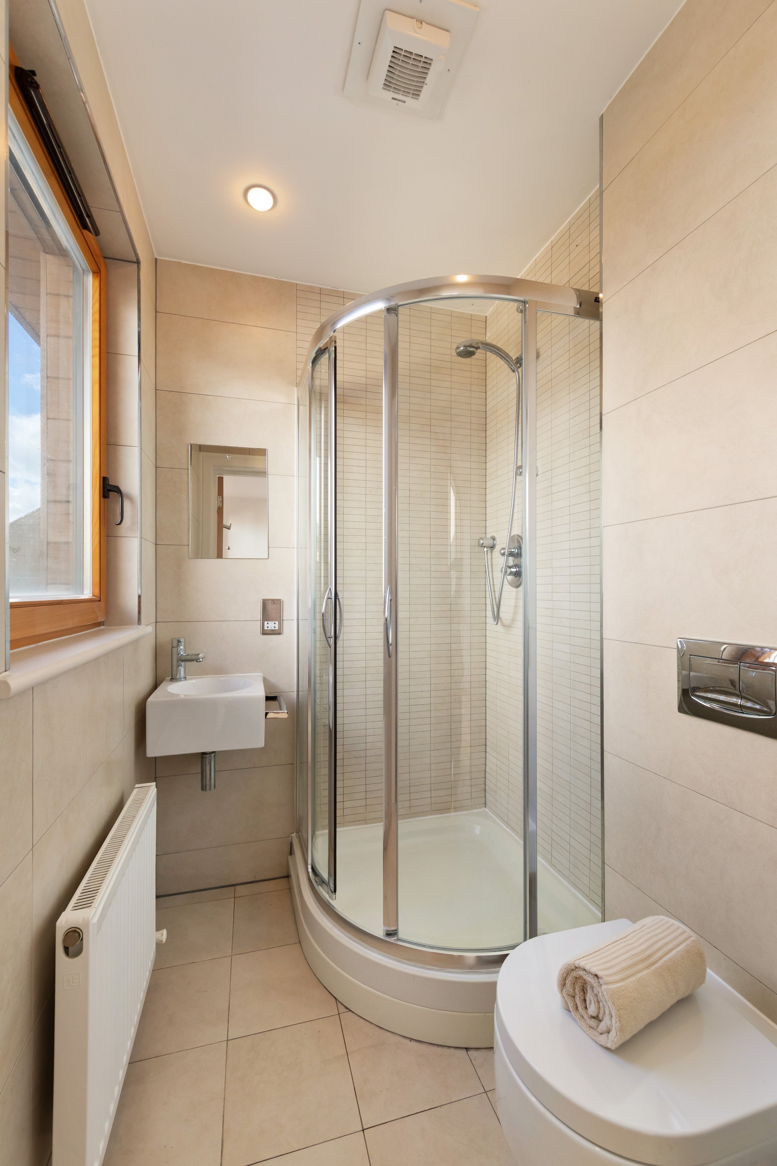 Small bathroom with a corner shower unit, a small wall-mounted sink, a window, a radiator, a mirror, and a towel on a round surface.
