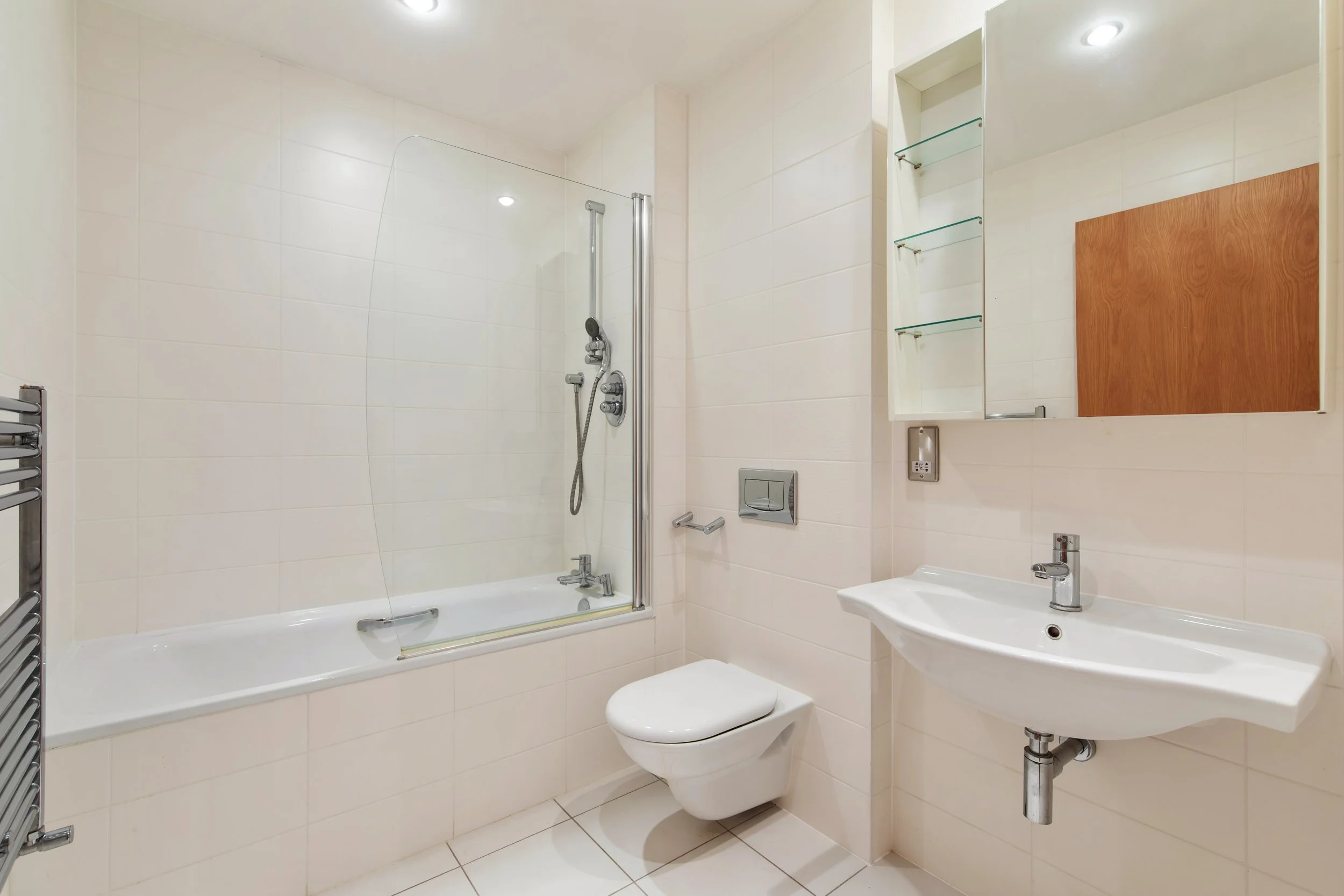 A modern bathroom with a white bathtub and a glass shower screen, a wall-mounted toilet, a white sink with a single lever faucet, a wall-mounted mirror cabinet with glass shelves, a heated towel rail, and beige tiled walls and floor.