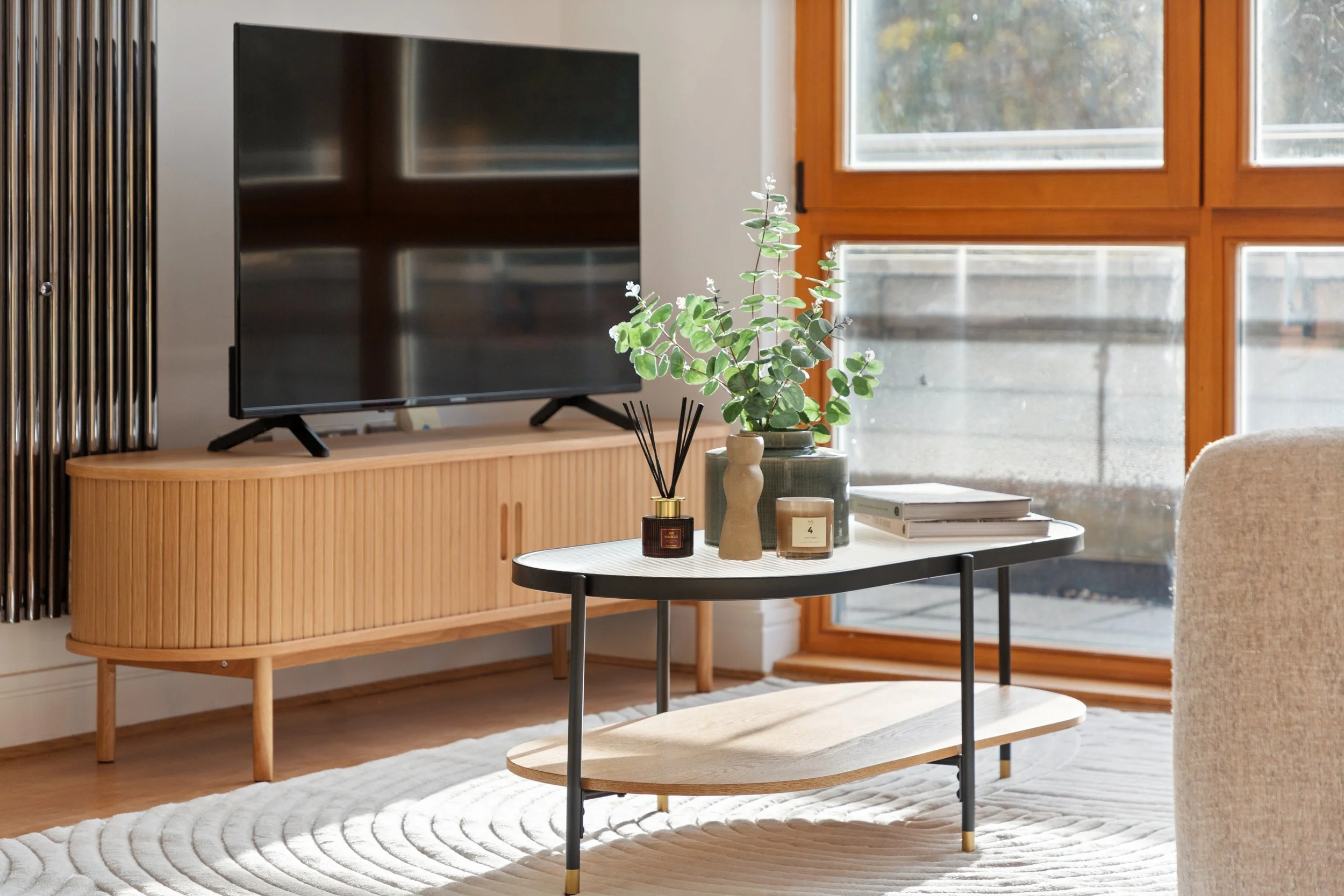 Modern living room with a wooden media console, a flat-screen TV, a round coffee table with decorative items, a large window with wooden framing, and a beige couch.
