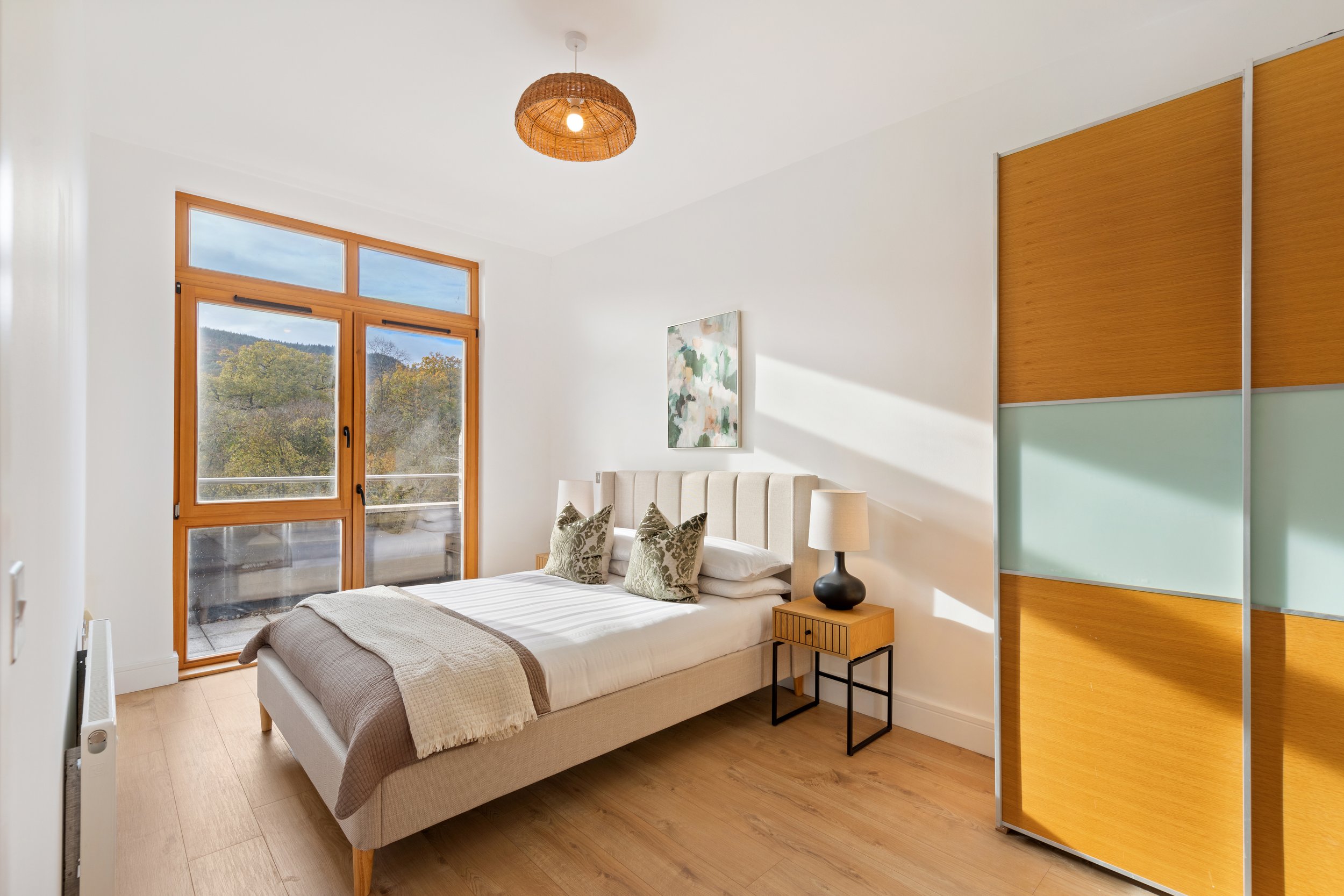 Minimalist bedroom with a large window showing trees and mountains, a beige bed with decorative pillows, a wooden nightstand with a black lamp, a framed artwork on the wall, and a wooden wardrobe with frosted glass panels.