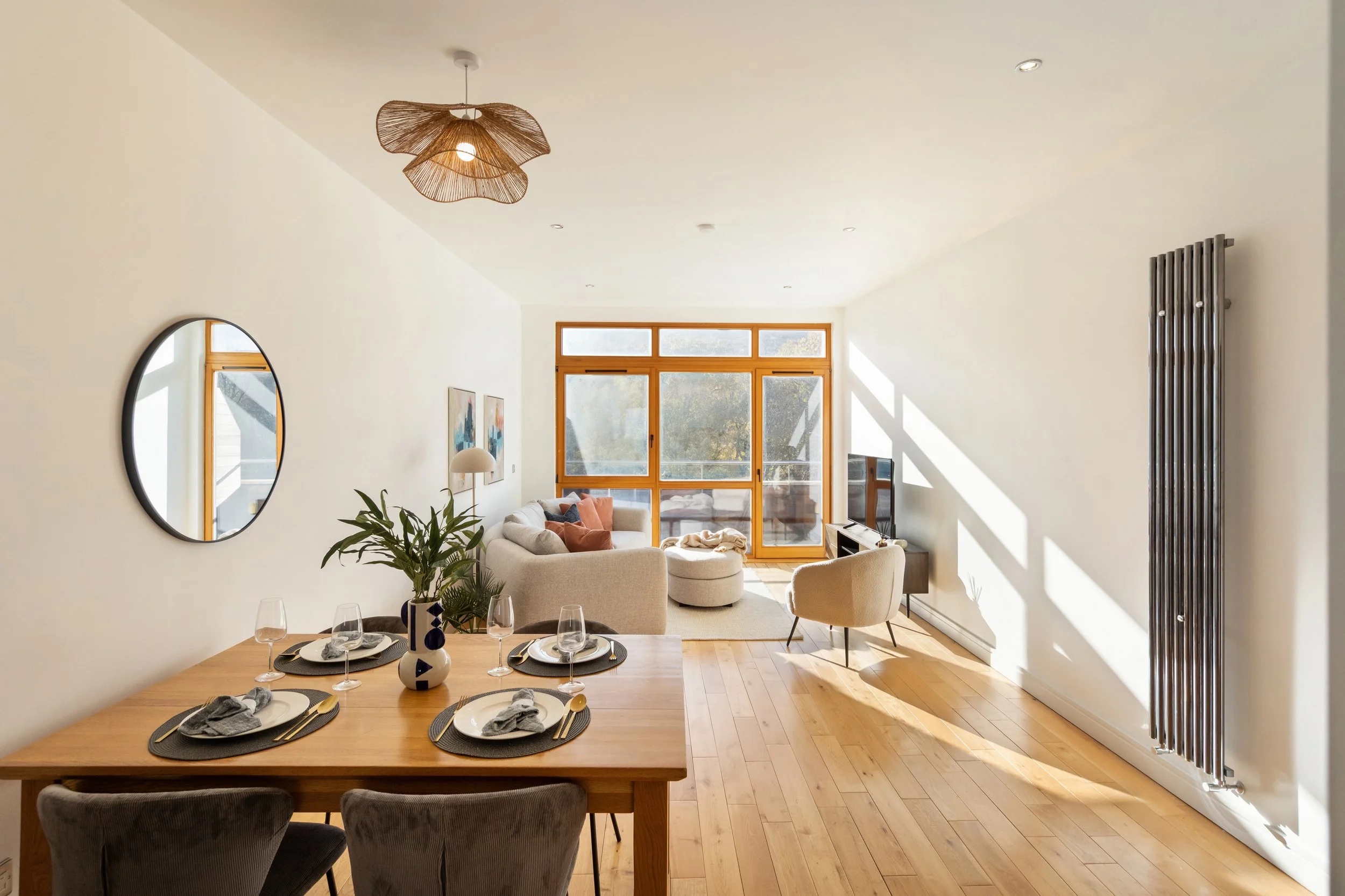 Open plan living and dining area with a wooden dining table, six place settings, gray chairs, a green plant, a white sofa with cushions, a cream armchair, a round ottoman, a television, large windows with wooden frames, and a wall-mounted radiator.
