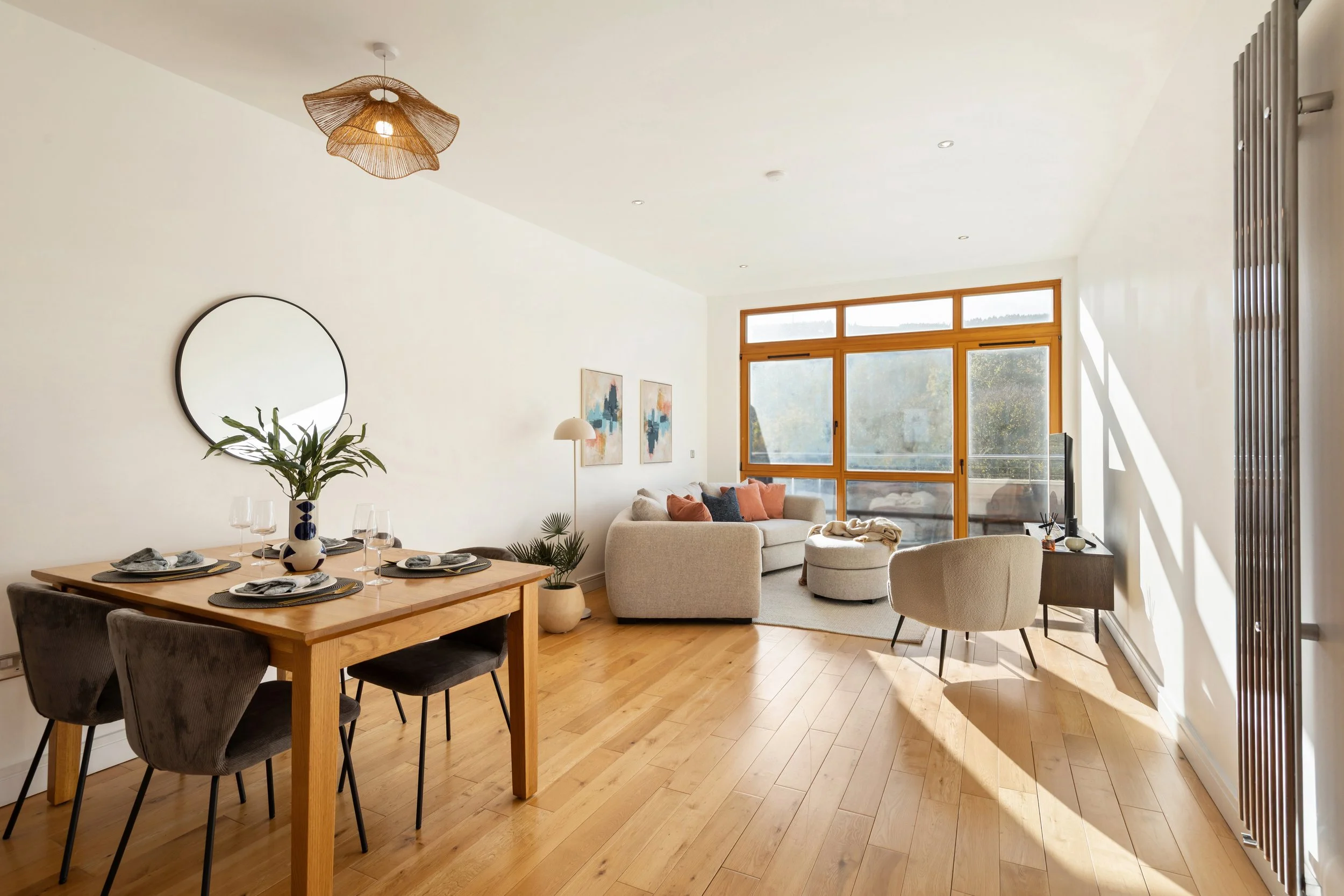 Living room with a dining area, beige sofa, armchair, large window, wooden flooring, wall art, and a modern ceiling light.