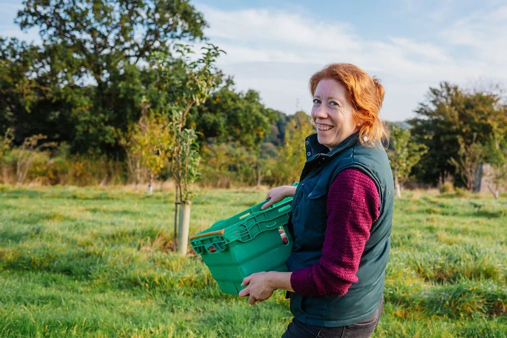 A woman with red hair holding a green basket, standing outdoors in a grassy area with trees.
