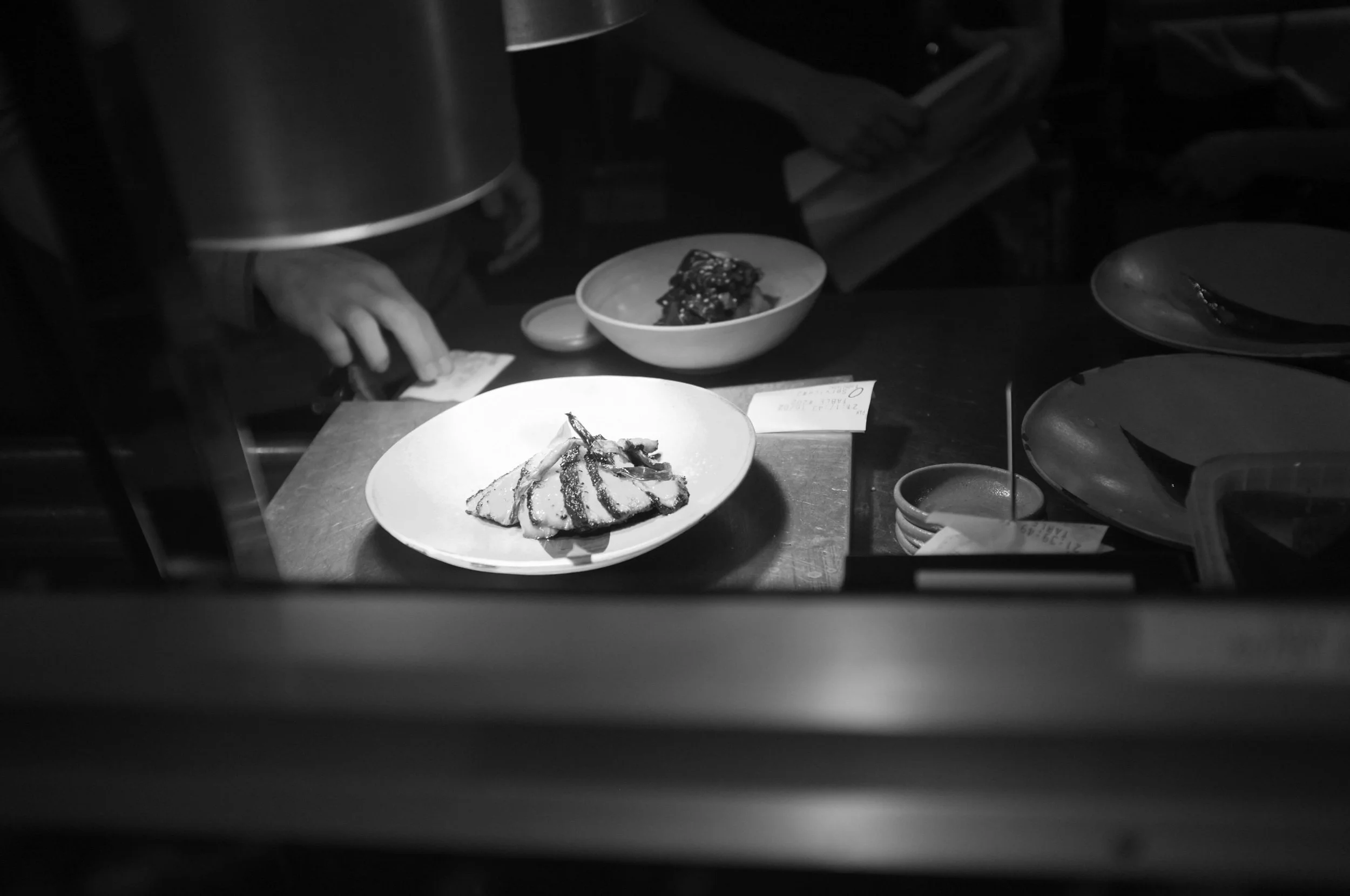 Black and white photograph of a restaurant kitchen with two plates of food on a counter, one with dessert and the other with a pastry or sandwich, with a person’s hand reaching for something in the background.