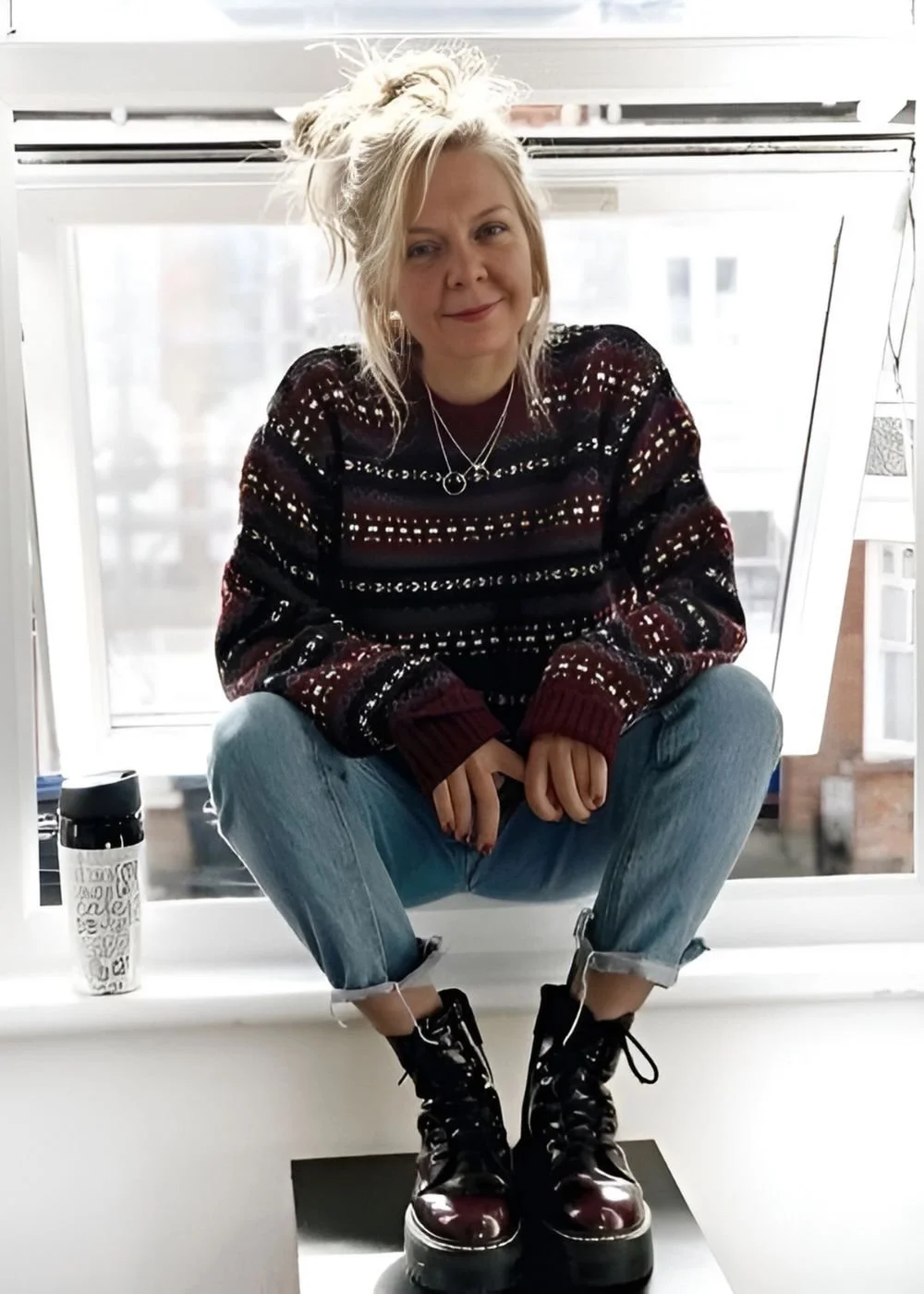 A woman with blonde hair tied up, wearing a patterned sweater, light blue jeans with rolled-up cuffs, and black combat boots, is squatting on a windowsill with a water bottle beside her, in front of a large window with a cityscape view.