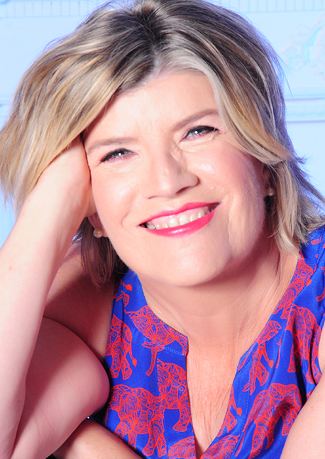 A woman with blonde hair smiling and resting her head on her hand, wearing a blue top with red patterns.