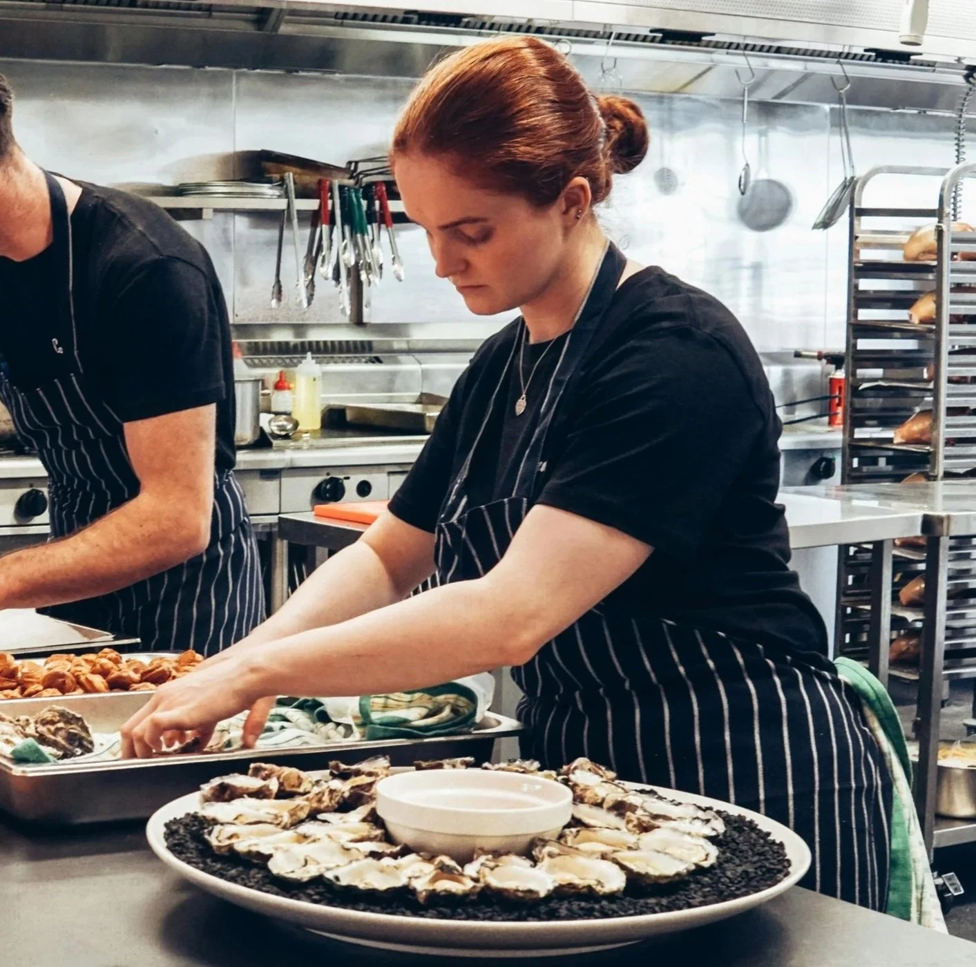 A woman with red hair, wearing a black shirt and striped apron, preparing oysters in a commercial kitchen.