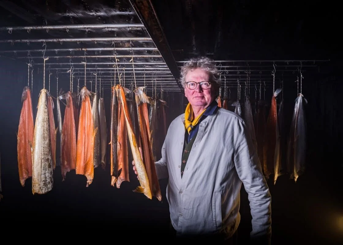 A man with glasses and curly gray hair standing in a smokehouse, surrounded by hanging smoked salmon fillets.