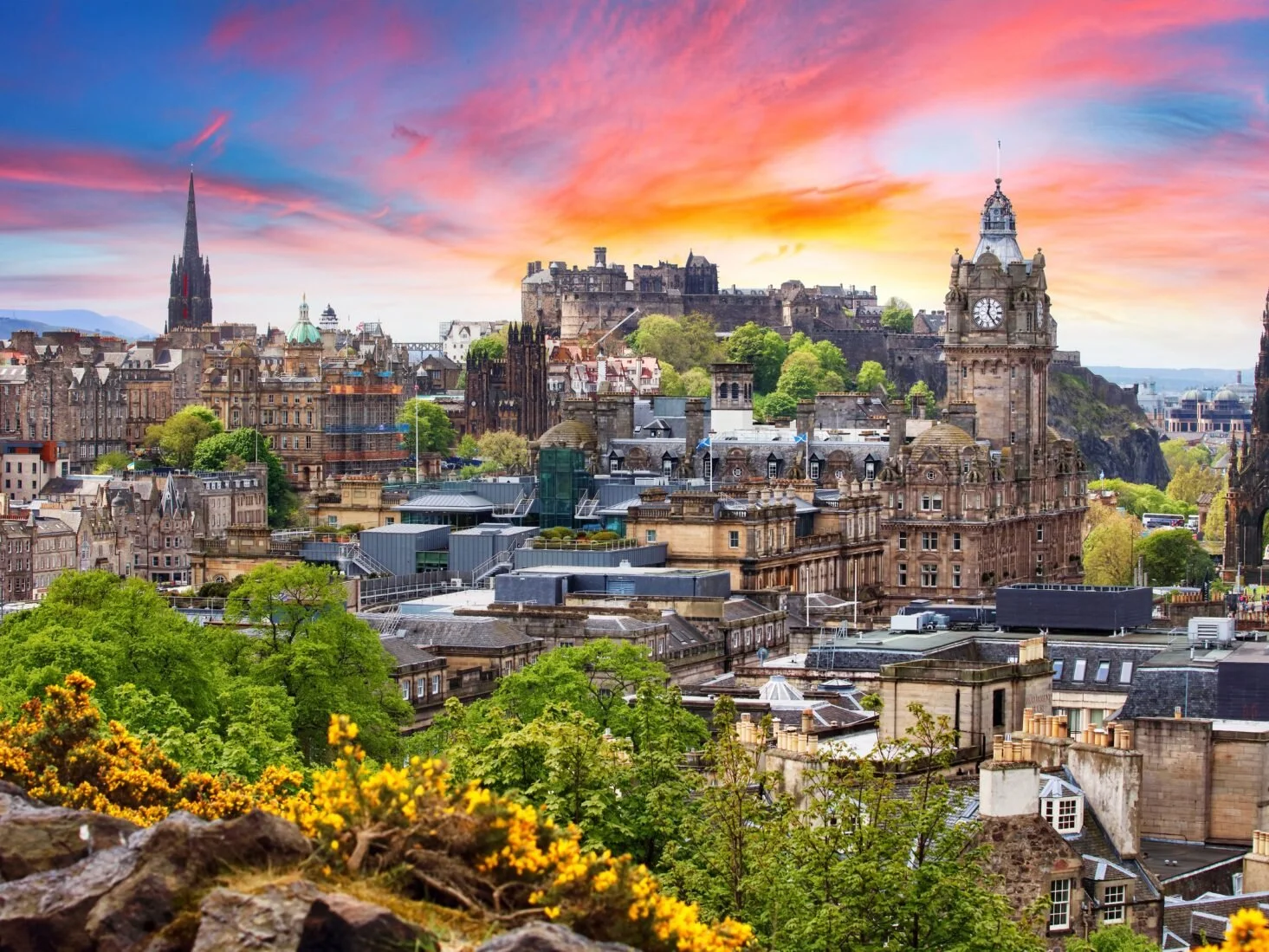 Sunset view of Edinburgh city skyline with historic buildings, a clock tower, and a castle on a hill in the background, with green trees and colorful flowers in the foreground.