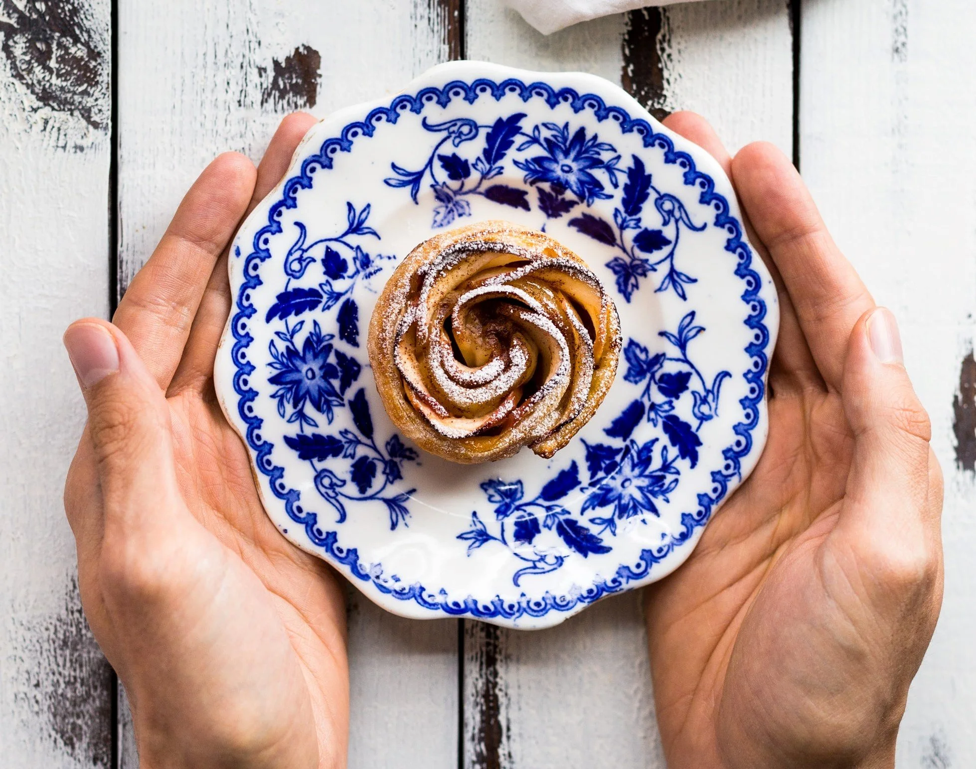 A person holding a blue and white floral patterned plate with a cinnamon roll sprinkled with powdered sugar on a white and brown wooden surface.