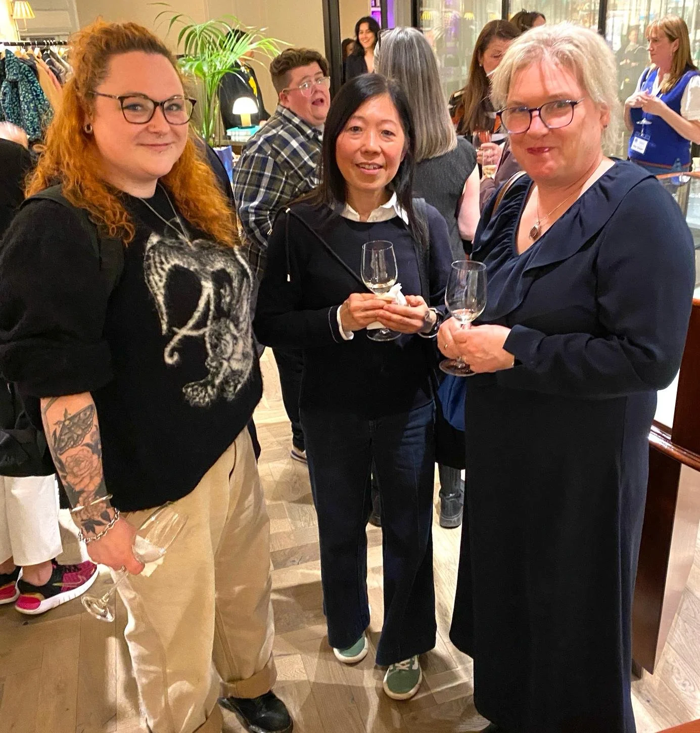 Three women holding wine glasses at an indoor social gathering, with a group of people in the background.