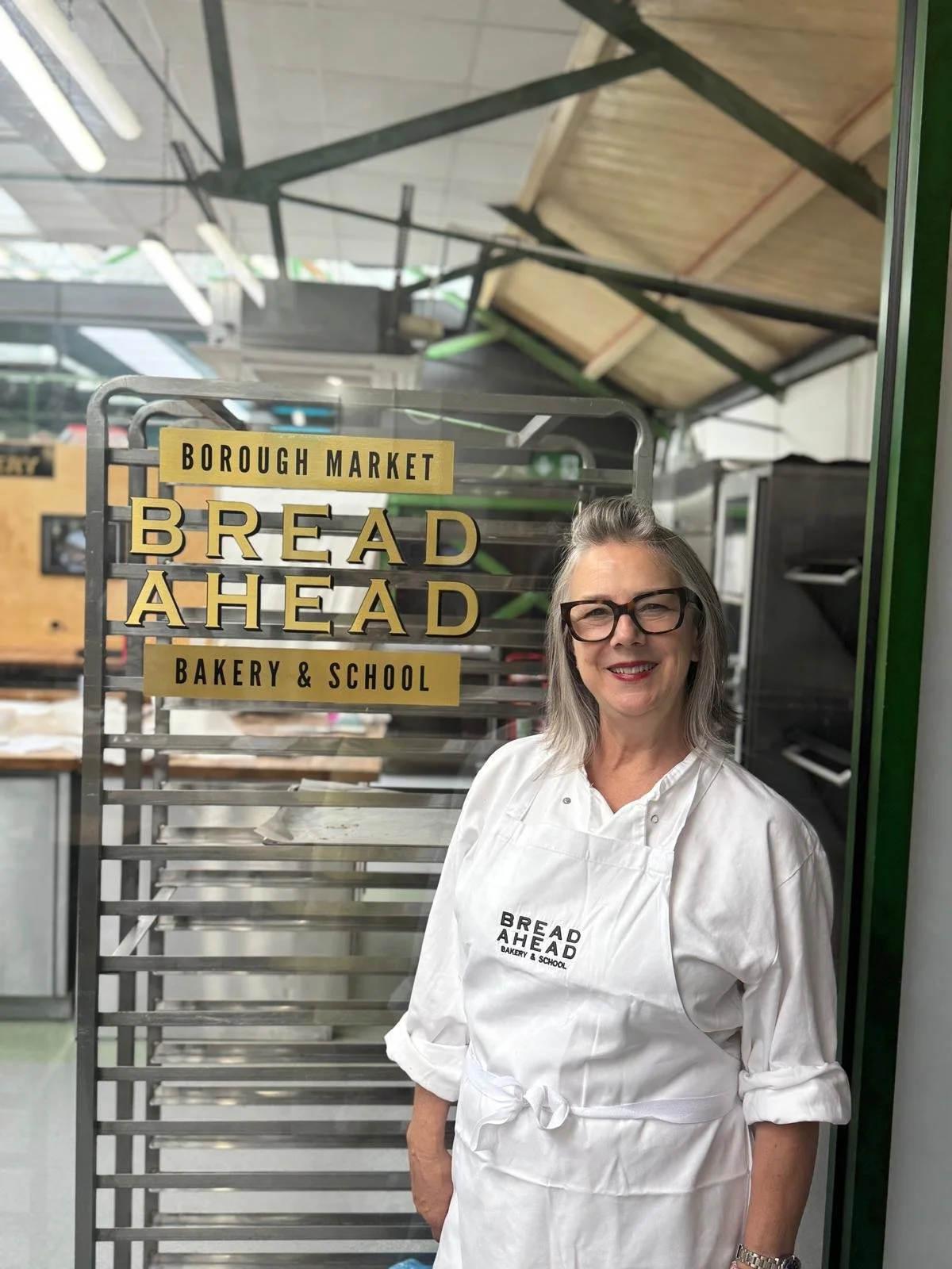 A woman with gray hair and glasses smiling, wearing a white apron with the words "BREAD AHEAD BAKERY & SCHOOL" printed on it, standing next to a bakery rack inside a bakery or market. There is a sign behind her that reads "Borough Market Bread Ahead Bakery & School".