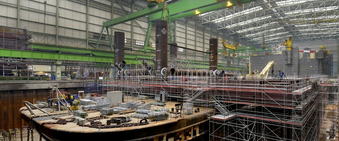 Inside a large shipyard with scaffolding surrounding a ship under construction, industrial equipment, cranes, and large pipes.