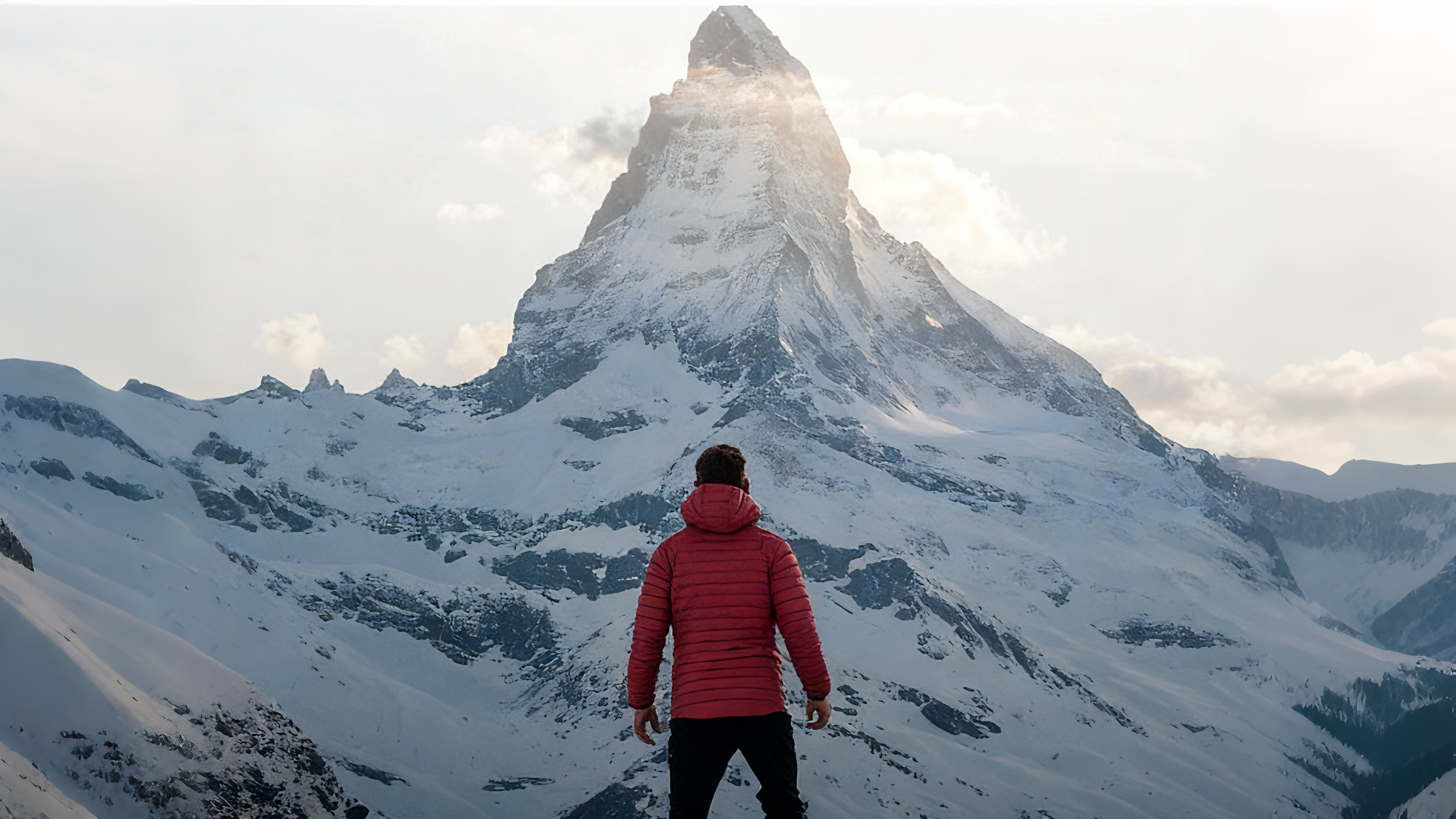 Person in a red jacket standing on snow-covered mountain landscape with the Matterhorn peak in the background.