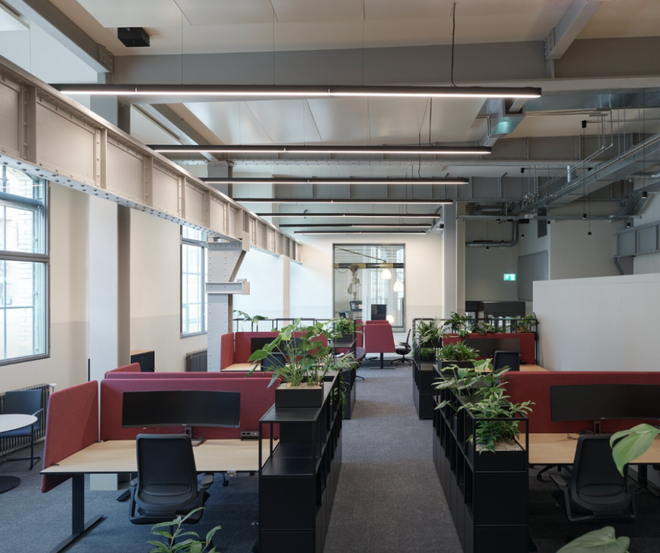 Modern office space with multiple workstations, red and black partitions, green potted plants, and natural light from windows.