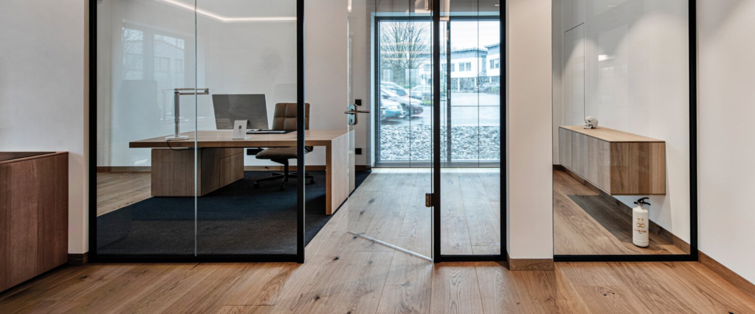 Modern office with glass walls, wooden furniture, and a view of the outside parking lot.