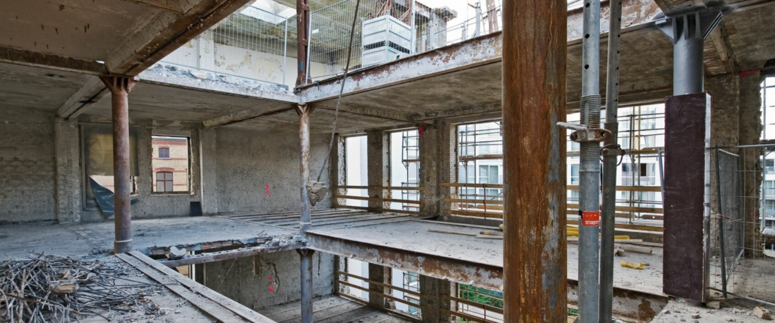 An unfinished building construction site with exposed concrete, metal support beams, scaffolding, and open spaces for windows, with construction materials and debris on the floor.