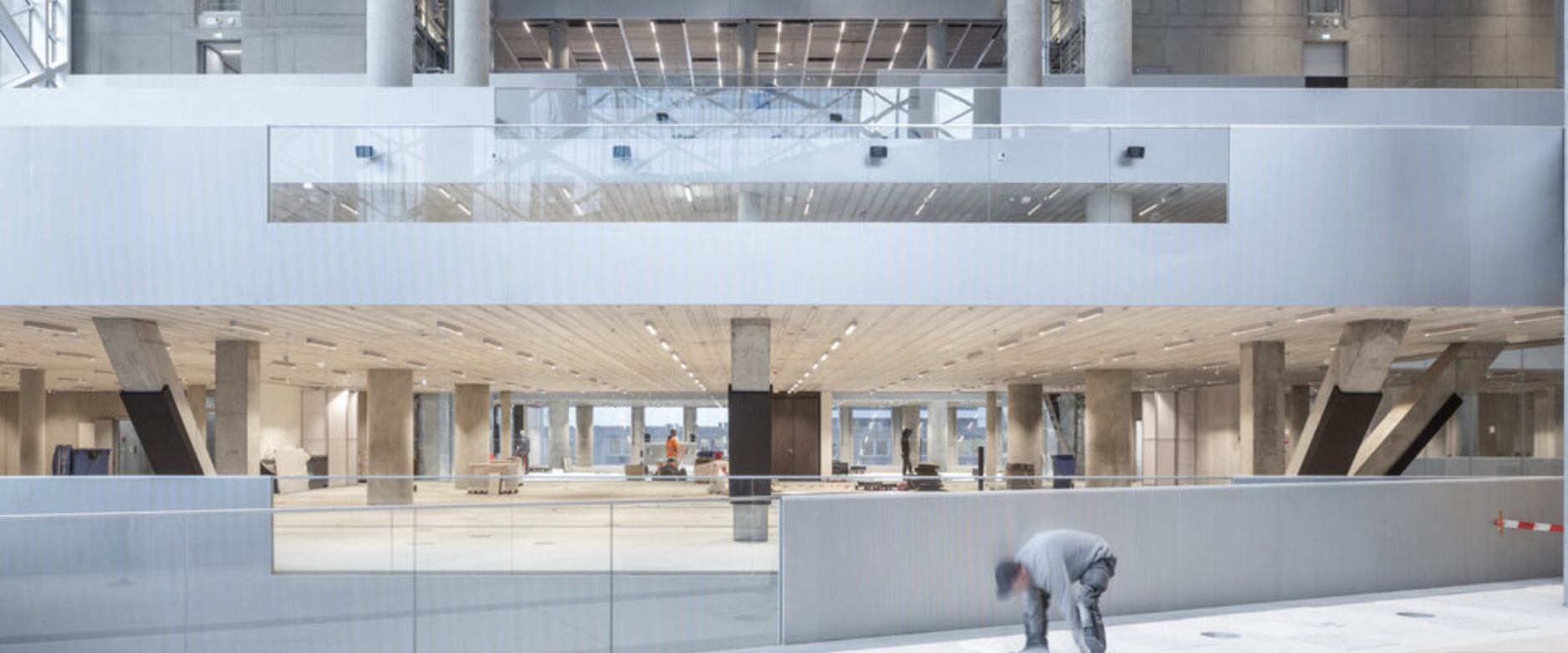Interior of a modern building under construction with multiple levels, concrete pillars, workers, and construction materials.
