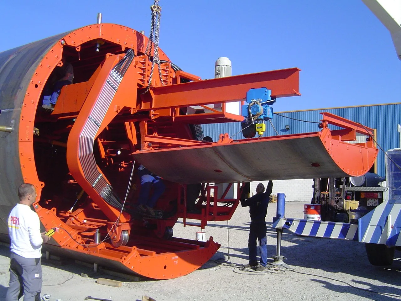 Workers assembling a large, orange spacecraft or missile on a ground platform outdoors, with a blue sky background.