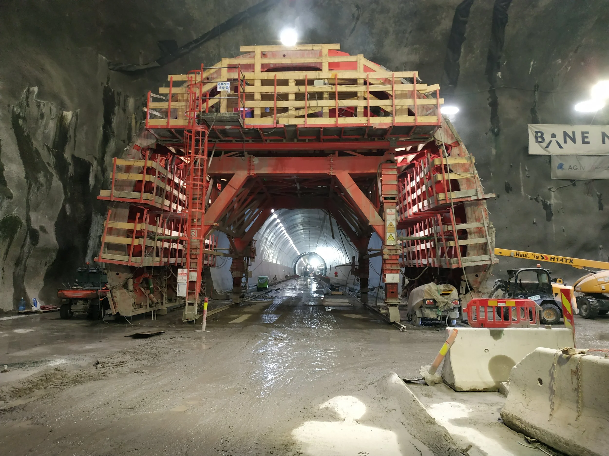 Underground tunnel construction site with large red scaffolding and machinery, wet ground, and an illuminated tunnel in the background.