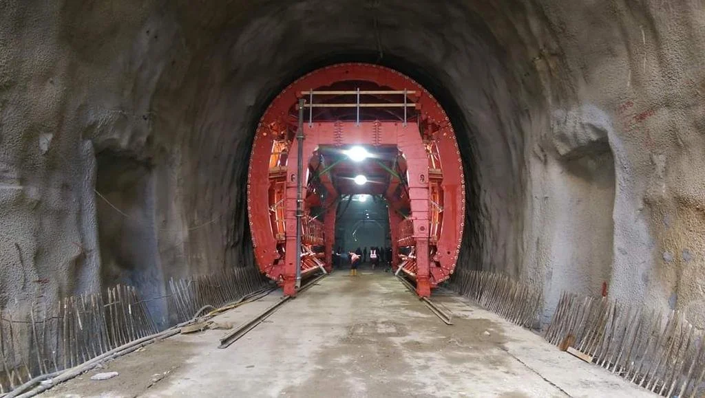 Construction workers and equipment inside a tunnel under construction, with a large red tunnel boring machine.