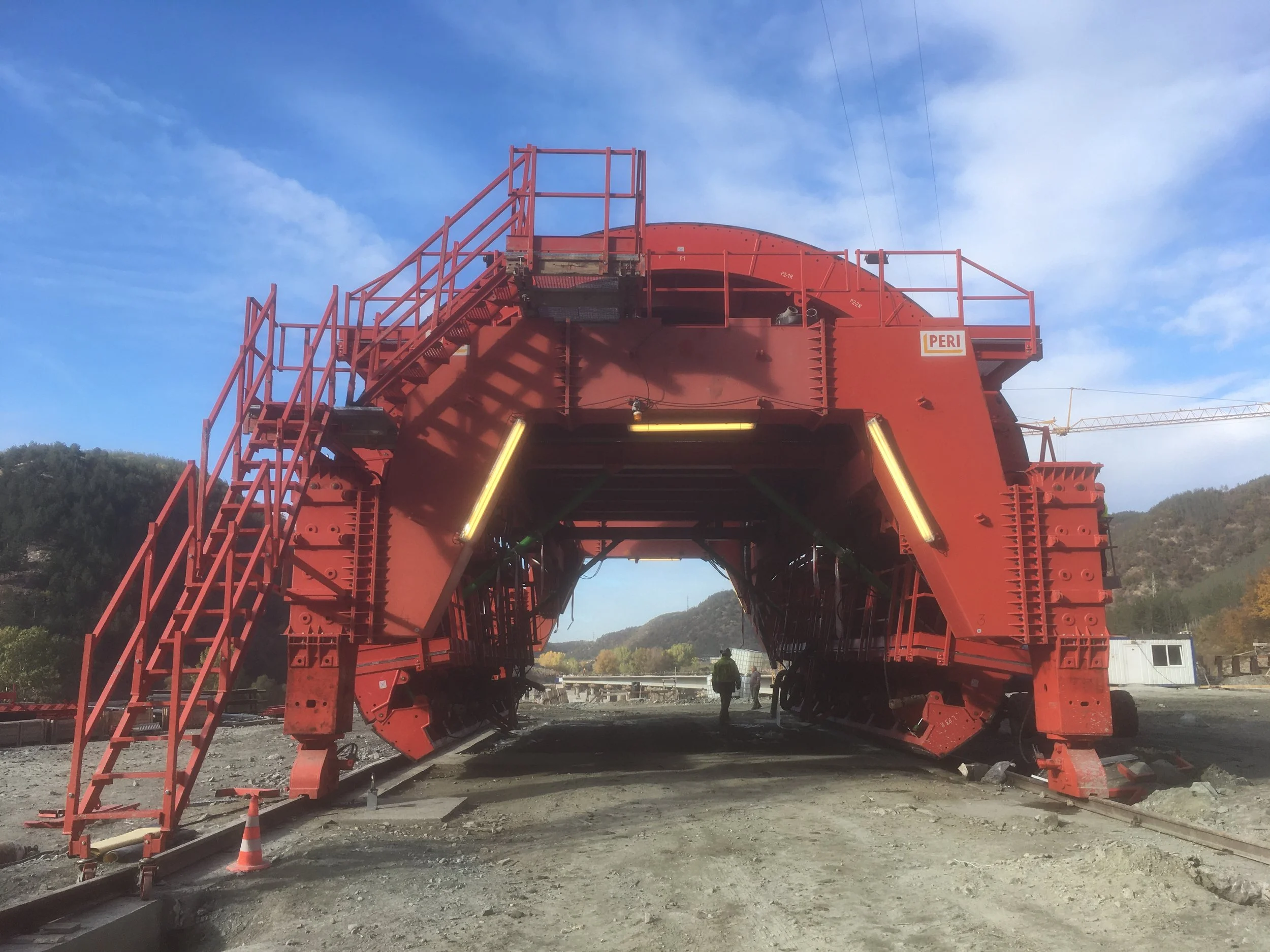 Large red train structure on railroad tracks with stairs on the left, under a partly cloudy sky with mountains in the background.