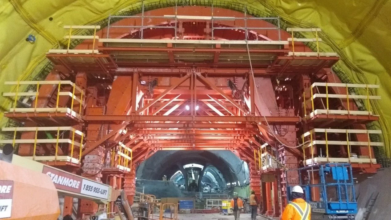 Inside a large tunnel construction site with tall scaffolding, workers in safety gear, and heavy equipment.