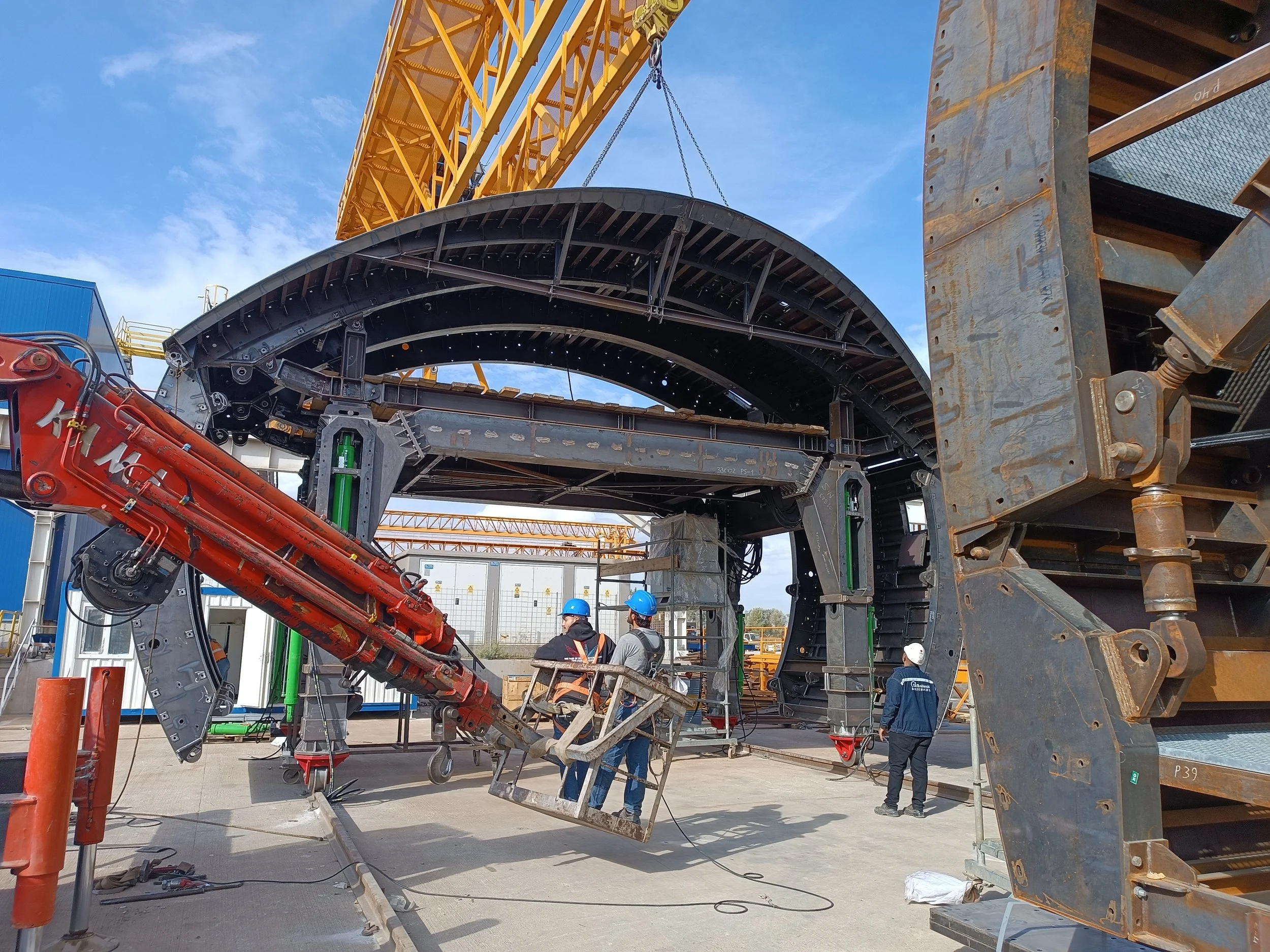 Construction workers in safety helmets working on a large steel structure with a yellow crane overhead, set against a bright blue sky.