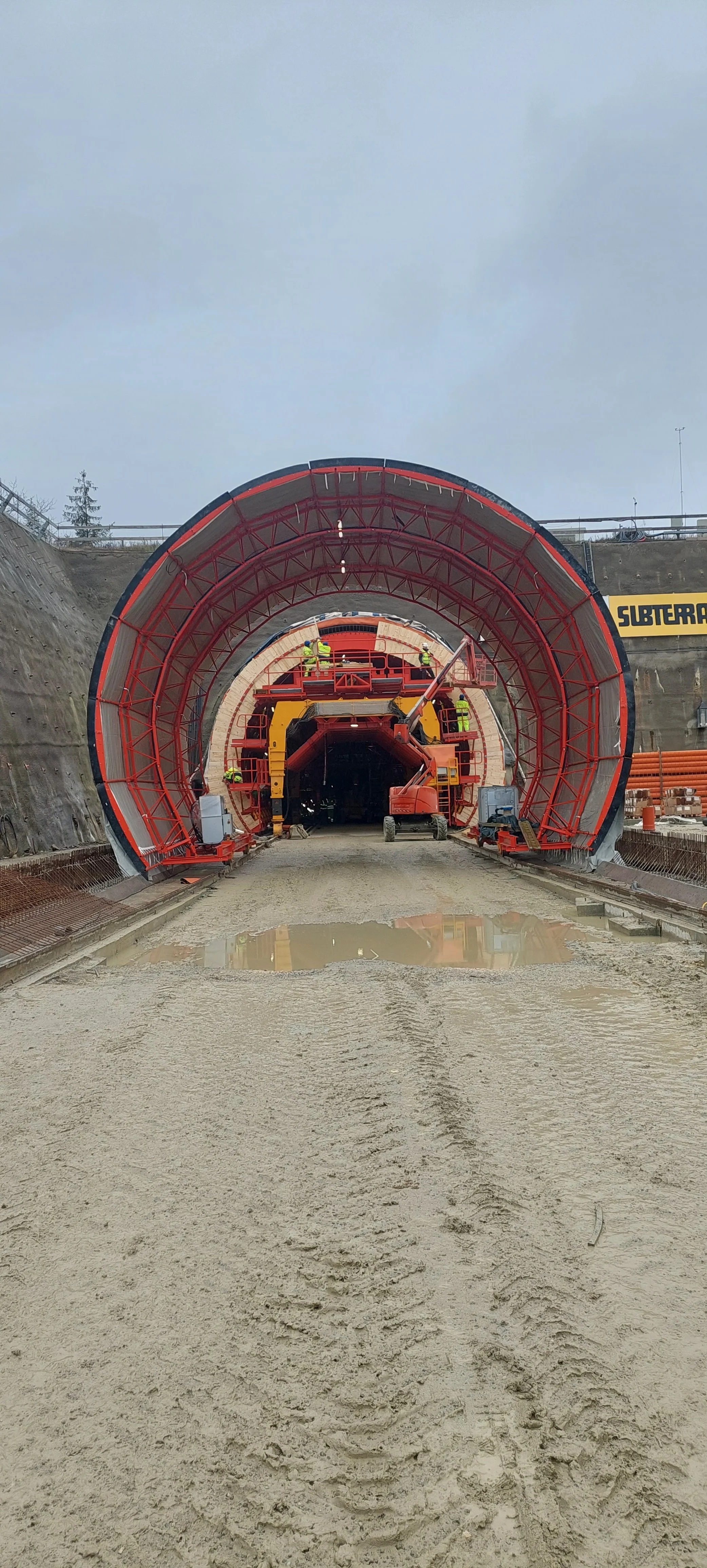 Construction workers and machinery working at a tunnel construction site with a partially built tunnel and wet, muddy ground in the foreground.