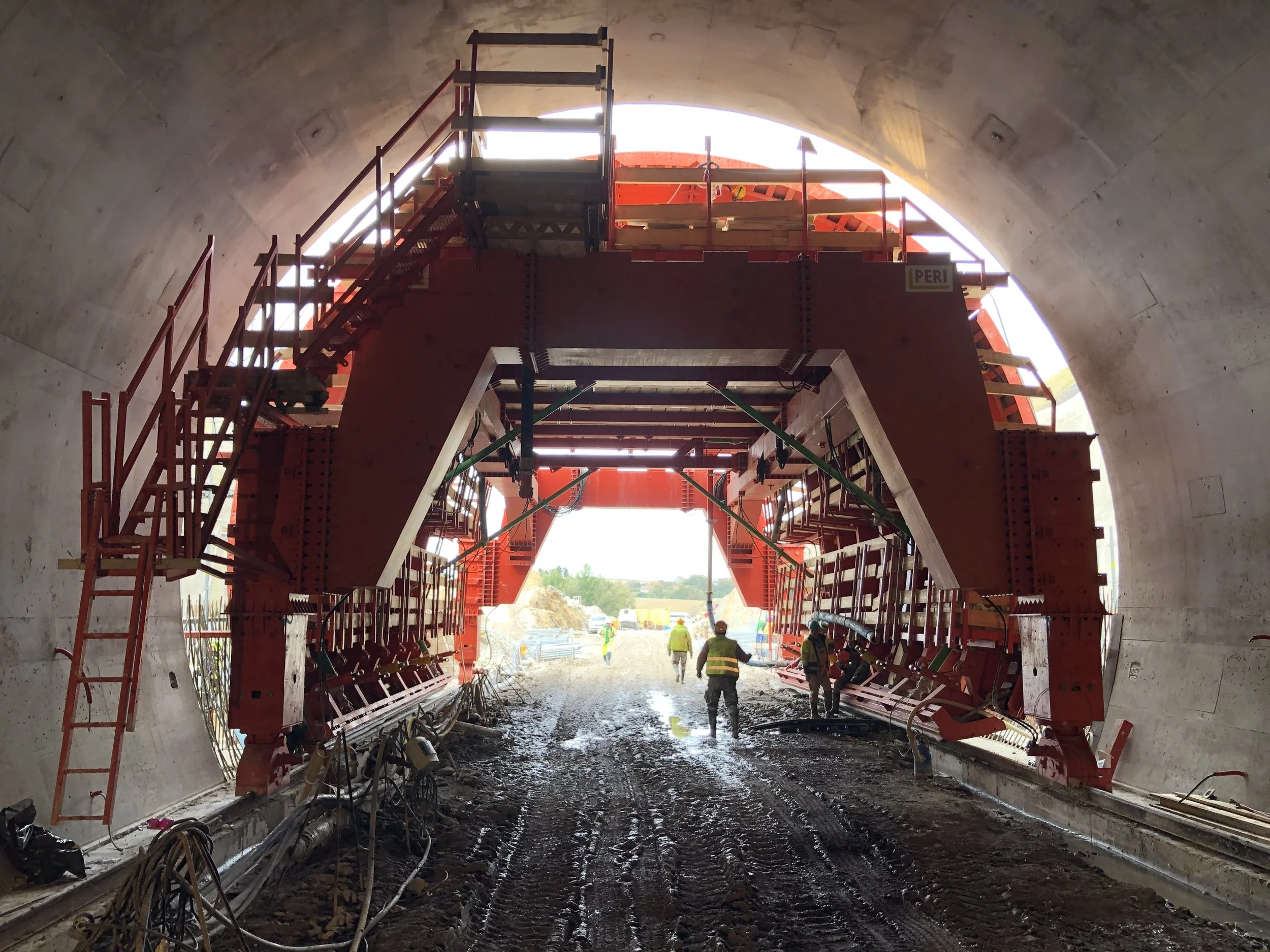 Construction workers inside a tunnel under construction, operating large red machinery to expand or reinforce the tunnel, with construction materials and muddy ground visible.