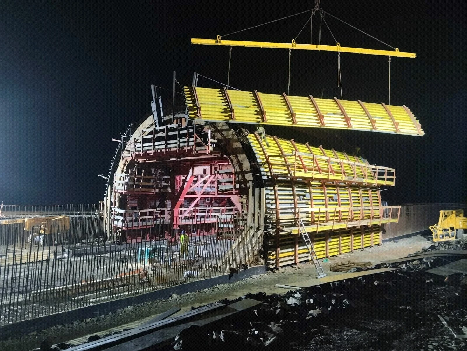 Construction workers and equipment work on a large tunnel boring machine frame at night, with the machine's yellow and red structural parts visible and overhead crane lifting a component.