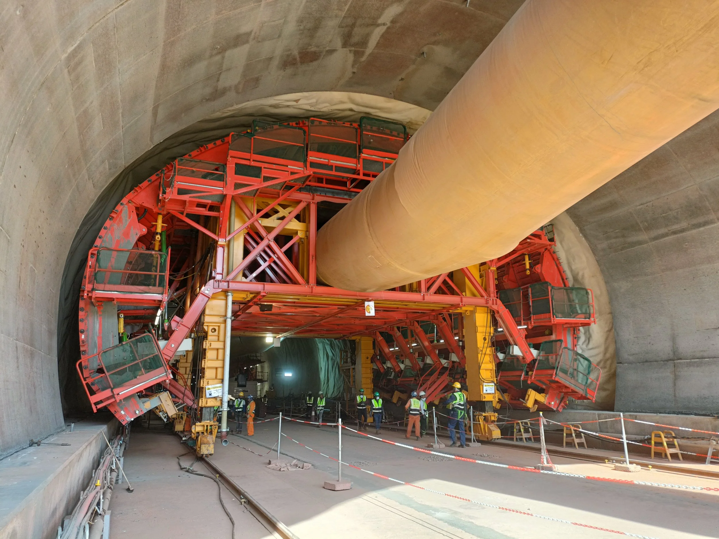 Construction workers are installing or maintaining a large tunnel support structure with a massive yellow pipe in a tunnel under construction.
