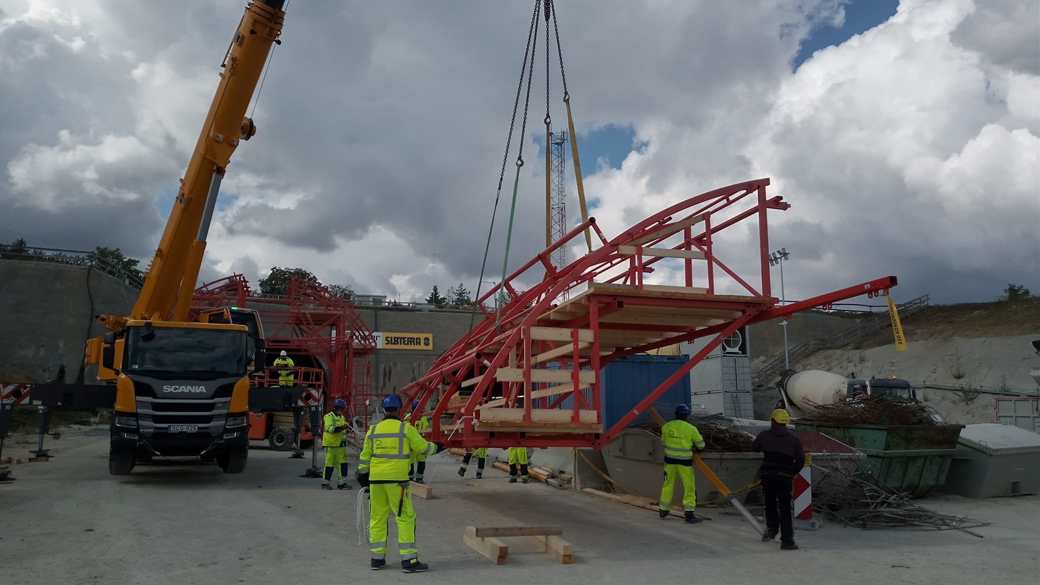 Construction site with workers and a large yellow crane lifting a red structural element.