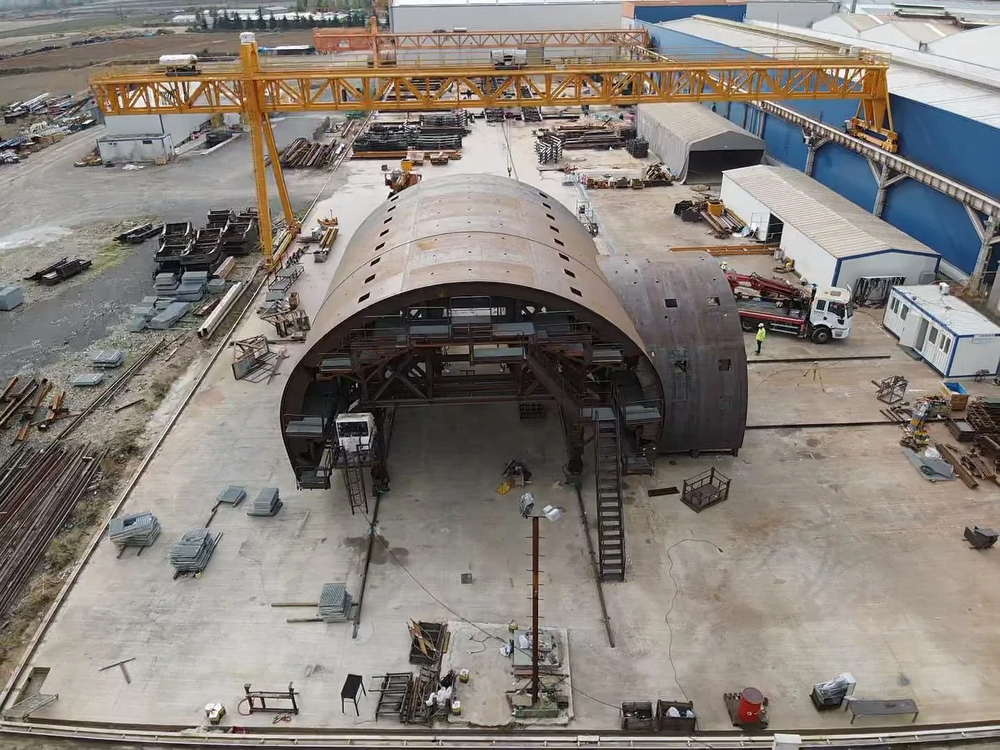 An industrial construction site with a large arched metal structure being assembled, surrounded by foundational work, equipment, and materials, with a tower crane overhead.