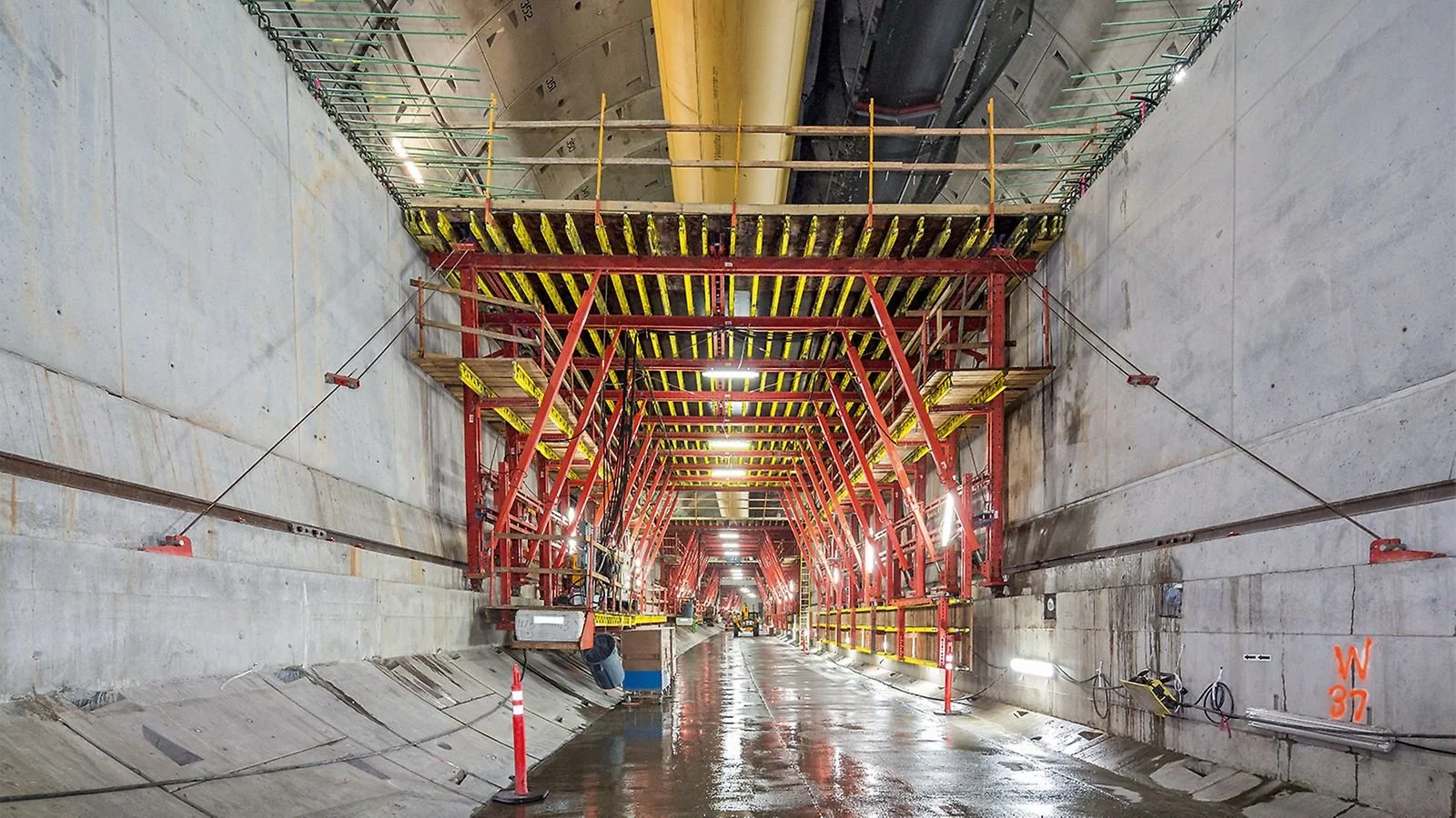 Underground tunnel construction site with red scaffolding and machinery, wet concrete floor, pipes, and bright lighting.