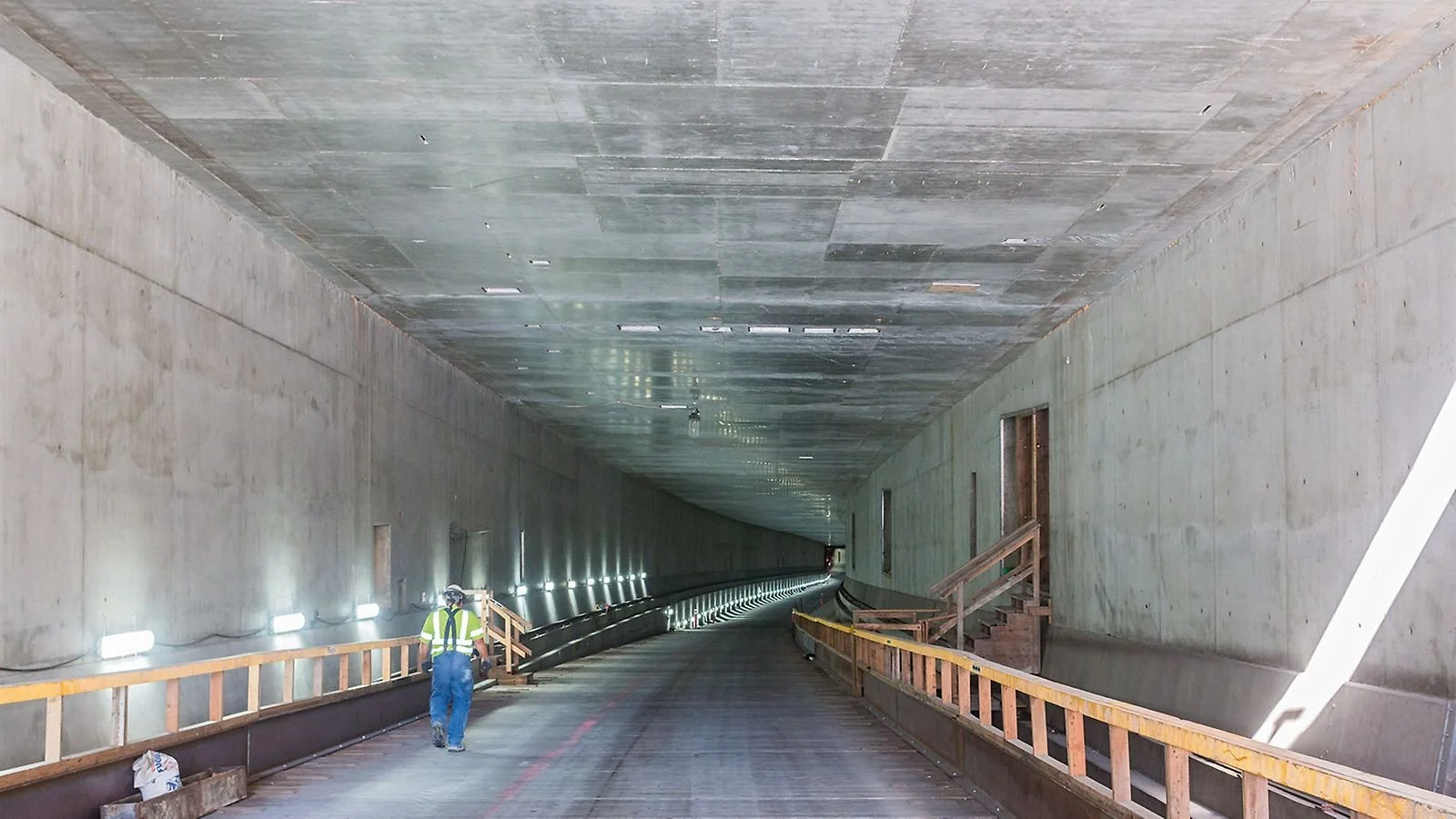 Construction worker walking inside a large tunnel with concrete walls and ceiling, illuminated by lights along the sides.